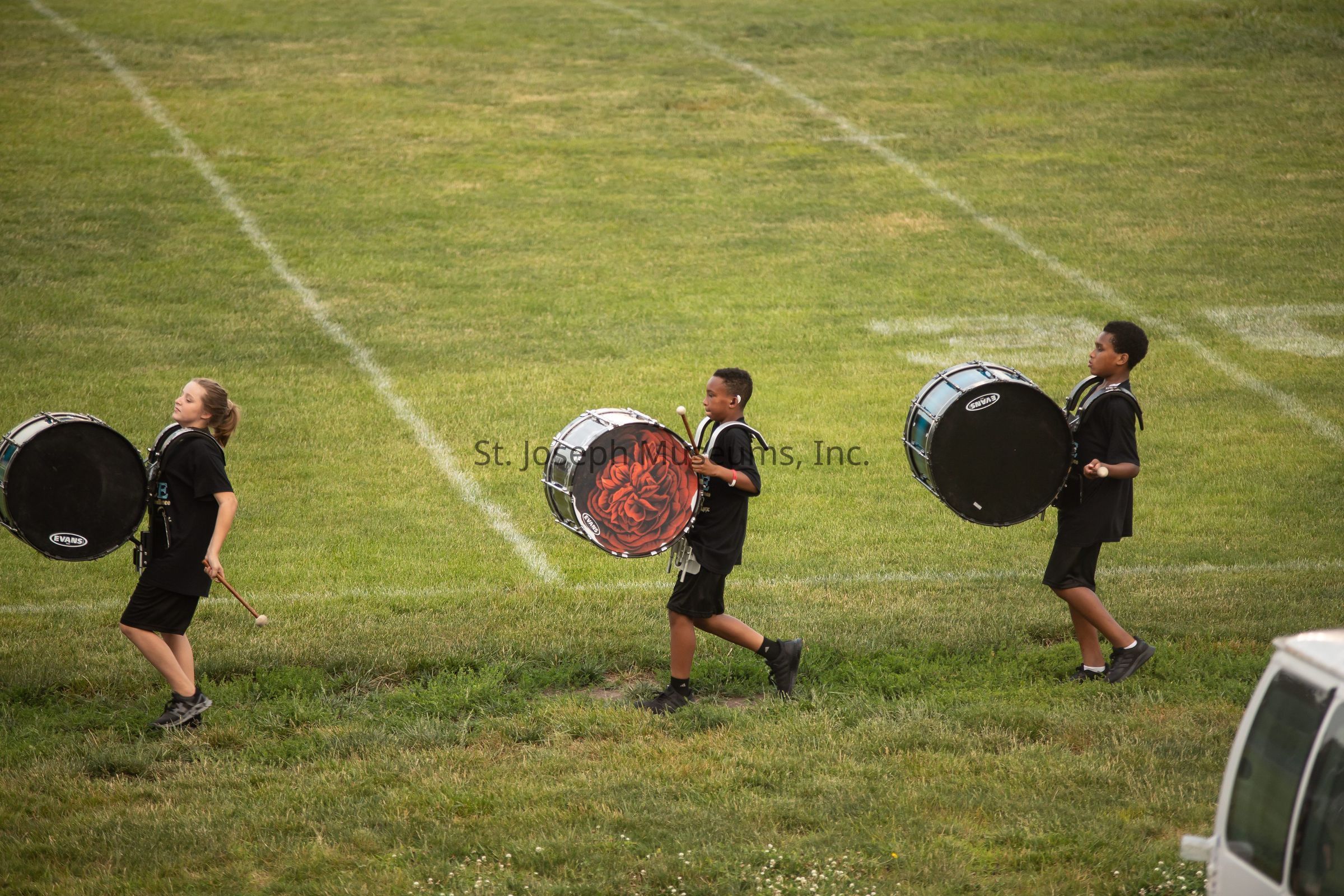 The image captures three young individuals marching across a grassy field, each carrying a large bass drum strapped to their bodies. They are aligned in a single file and appear to be part of a marching band, engaging in what seems to be a rehearsal or performance. The first individual, holding drumsticks, leads the group, followed closely by the others, each drum featuring a unique design, with one drum notably adorned with a vivid red pattern resembling a flower. They all wear coordinated outfits, suggestive of a team or school event, moving with synchronized precision. Their expressions and posture convey concentration and rhythm, emphasizing their commitment to the musical performance. The expansive background consists of well-maintained green grass marked with faint white lines, possibly indicating a sports field or similar open space, providing a serene yet dynamic setting for the musicians. The image exudes a sense of youthful energy, teamwork, and the joy of participating in collective artistic endeavors, highlighting the discipline and dedication involved in such group activities.