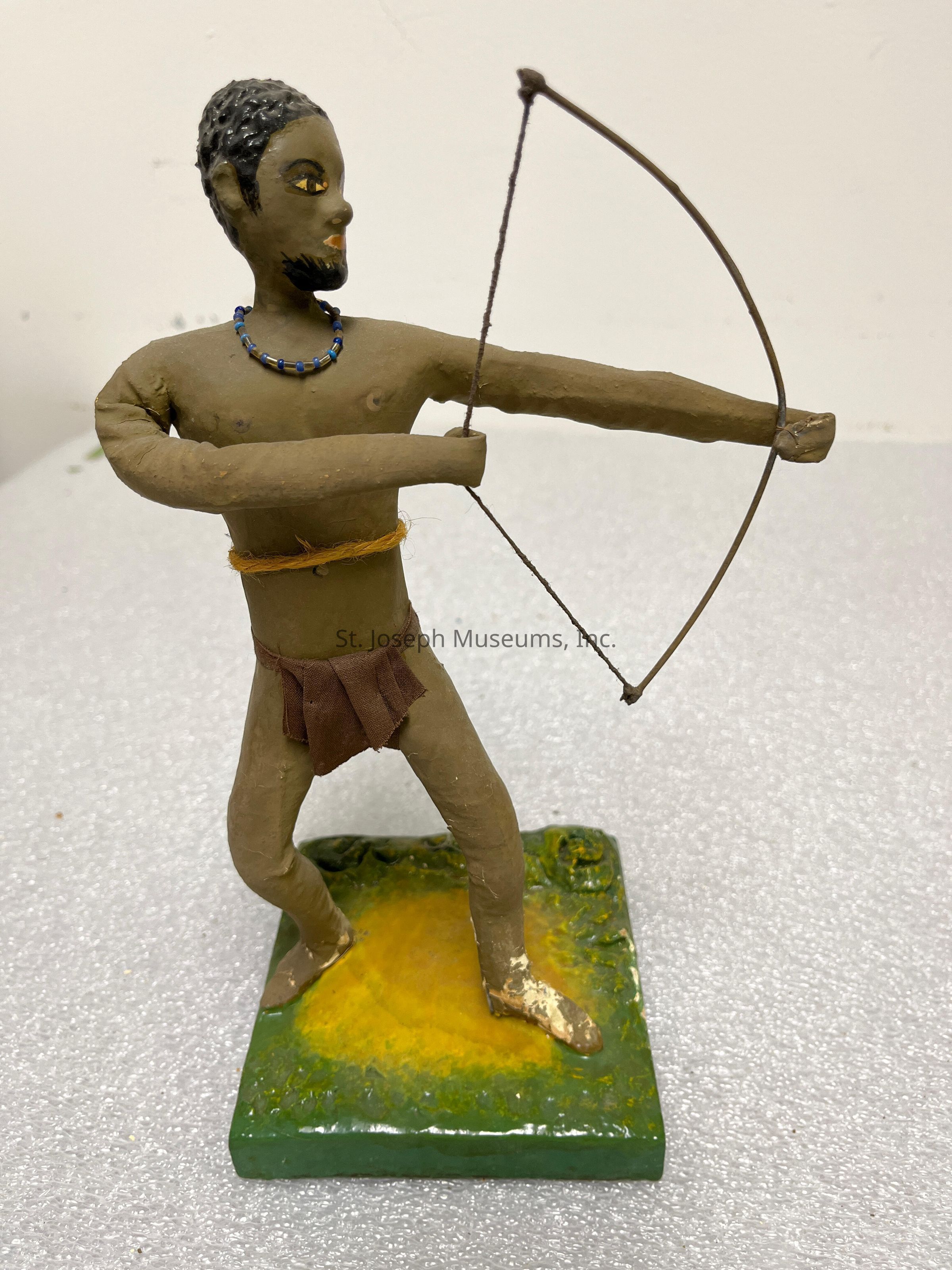 The image depicts a crafted figurine of a person with a bow and arrow, poised as if preparing to shoot. The figure stands on a square base with a textured surface, painted in green and yellow hues to suggest grass or earth. The figurine is fashioned with prominent facial features, including painted eyes and a beard, adding a lifelike touch. It wears a simple brown loincloth and a blue beaded necklace. The string of the bow is taut, illustrating a sense of motion and readiness, while the person’s stance, with one leg slightly forward, demonstrates balance and focus. Each limb is intricately shaped to reflect a natural human form.
