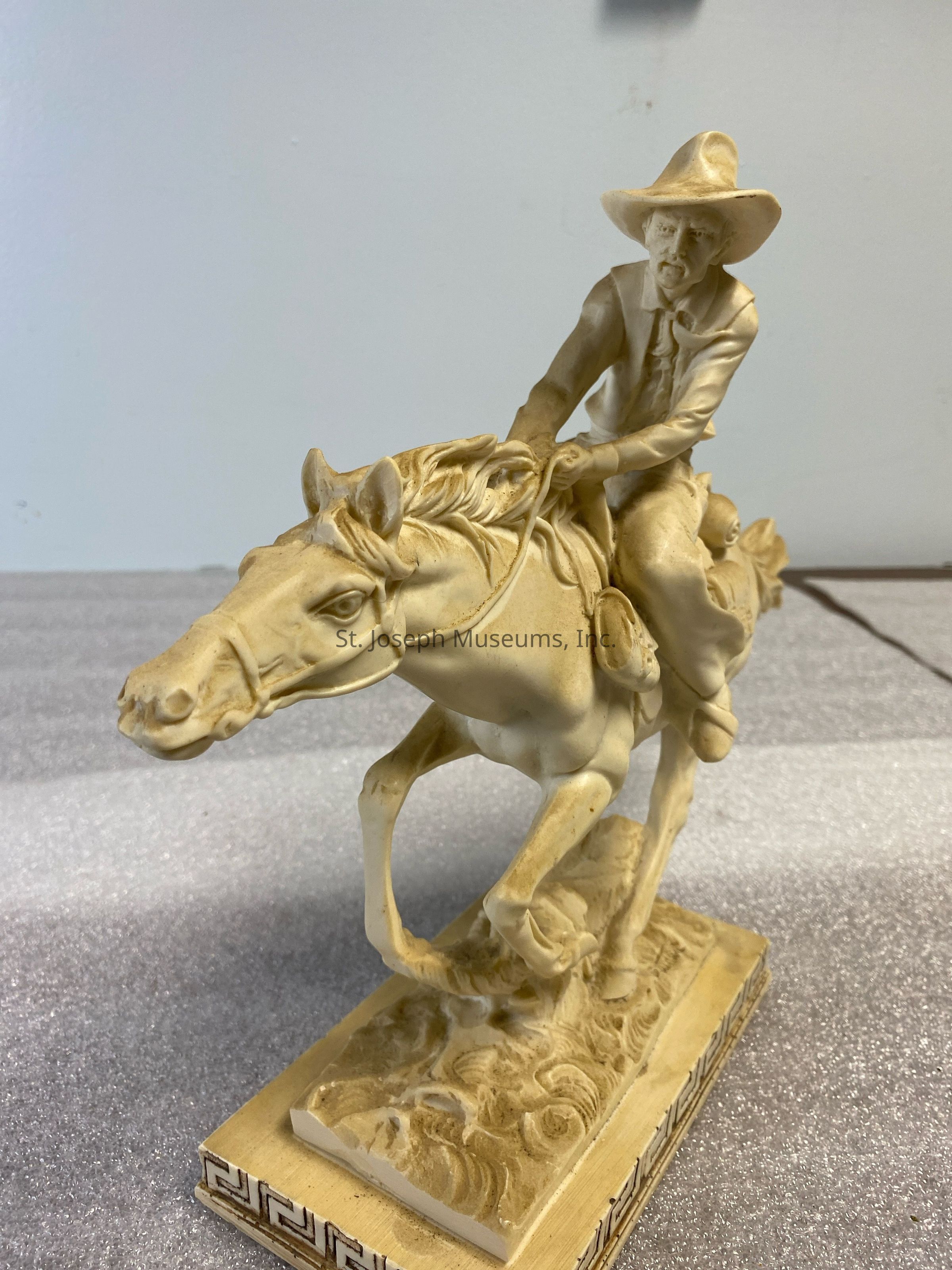 The image depicts a detailed ivory-colored sculpture of a cowboy in a dynamic pose, riding a galloping horse with a focused expression as he holds the reins tightly, capturing a sense of movement and adventure; the base of the sculpture features intricate patterns and textures, enhancing its artistic appeal and evoking a sense of the wild frontier.