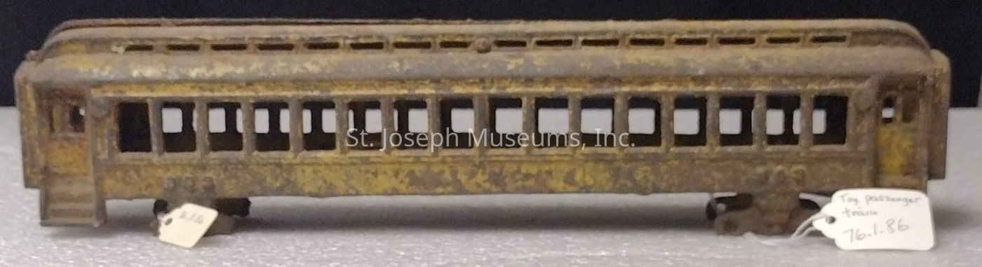 The image depicts an old, rusted toy train car, primarily in a yellowish hue, resting on a flat surface. The toy is designed to resemble a passenger train car, featuring a series of evenly spaced rectangular windows along its sides. Despite its weathered appearance, suggesting it has been exposed to the elements or undergone significant aging, the train retains a distinct and detailed design. The toy rests on small wheels, and beneath it are two handwritten tags: one on the left appears to show a number, and the other on the right reads "Toy passenger train 76.1.86," indicating it might be part of a collection or catalogued item.