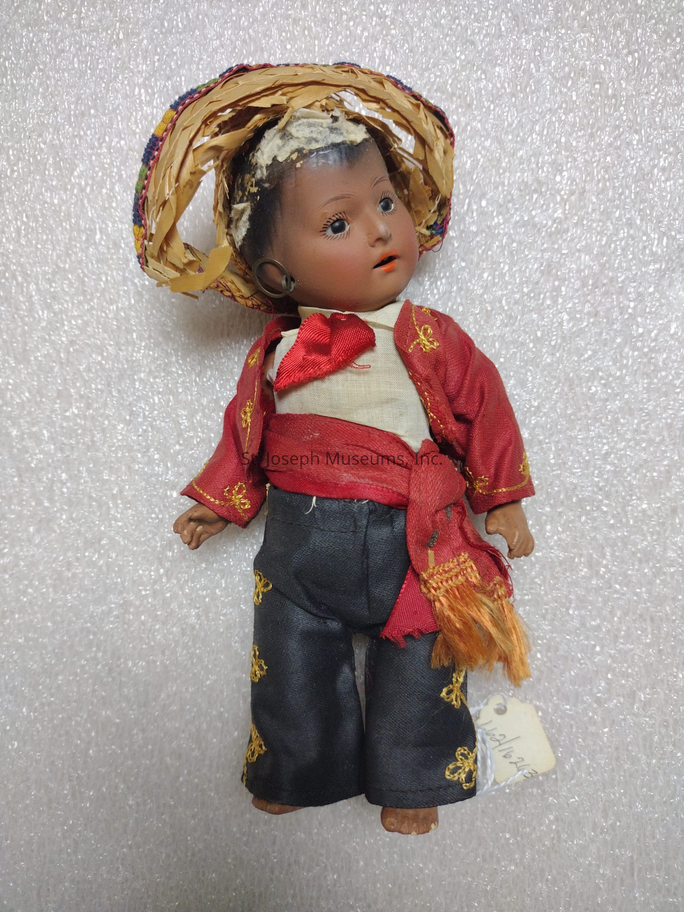 The image shows a vintage doll dressed in traditional attire, featuring a colorful woven hat on its head and a red jacket adorned with decorative yellow patterns over a white shirt. The doll is also wearing black pants with similar yellow embroidery and a red sash tied around its waist. The facial expression of the doll is neutral, with distinct eyes that are open, giving it a lifelike appearance. The clothing, although aged, still reflects intricate craftsmanship, and there is a tag attached to the doll, though the details on it are not fully visible. The background is a textured white surface, enhancing the doll's vibrant colors and intricate details.
