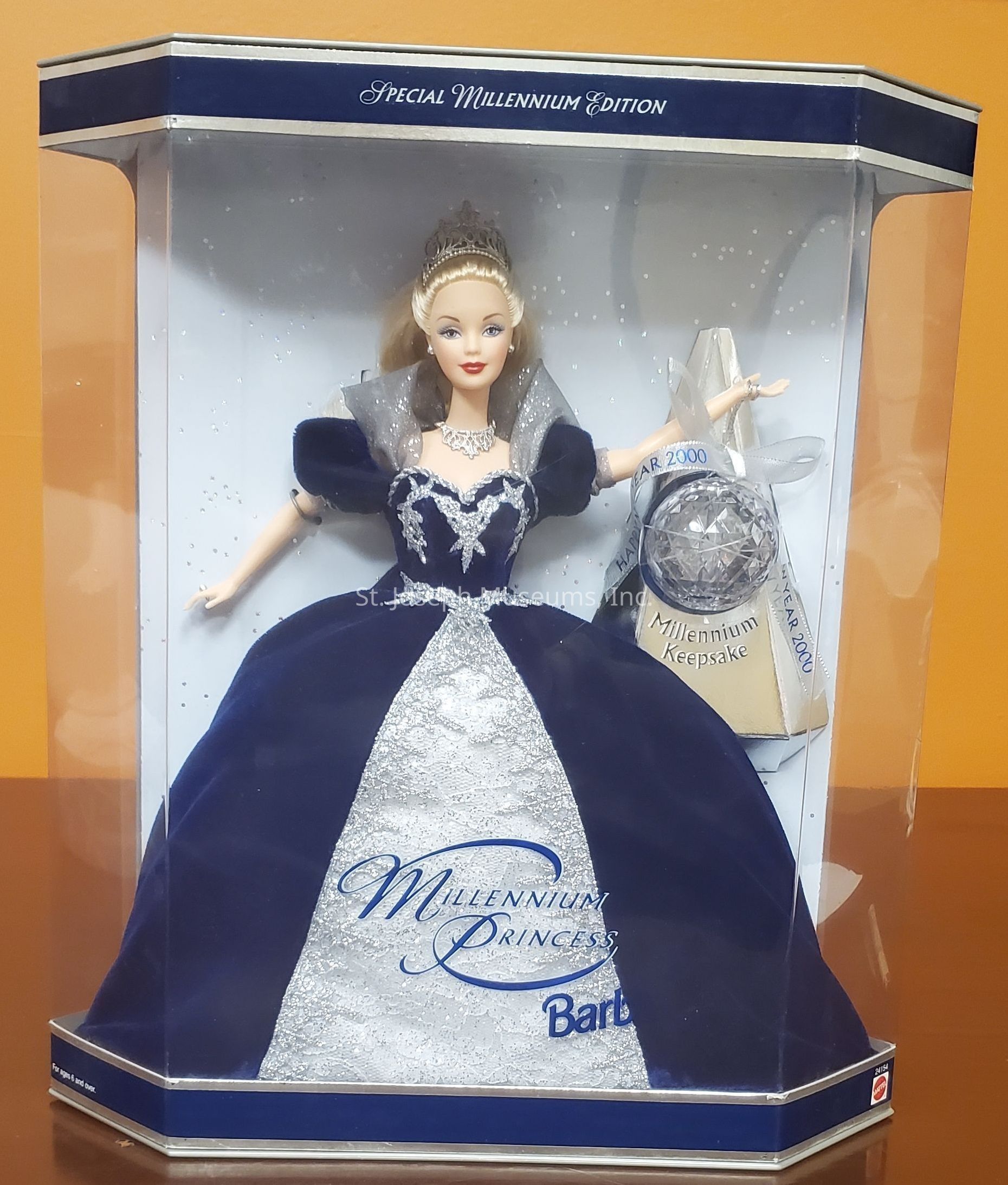 The image displays a "Special Millennium Edition" Barbie doll, elegantly enclosed in a transparent packaging, showcasing the "Millennium Princess" theme. The doll is adorned in a luxurious deep blue gown accented with silver, featuring intricate silver detailing across the bodice and skirt. She wears a sparkling tiara and a necklace, adding a touch of regal charm. Beside her, an ornamental silver keepsake, labeled "Millennium Keepsake," inscribed with "Year 2000," is carefully positioned. The packaging is designed with navy blue borders, emphasizing the special edition status and commemorating the turn of the millennium.