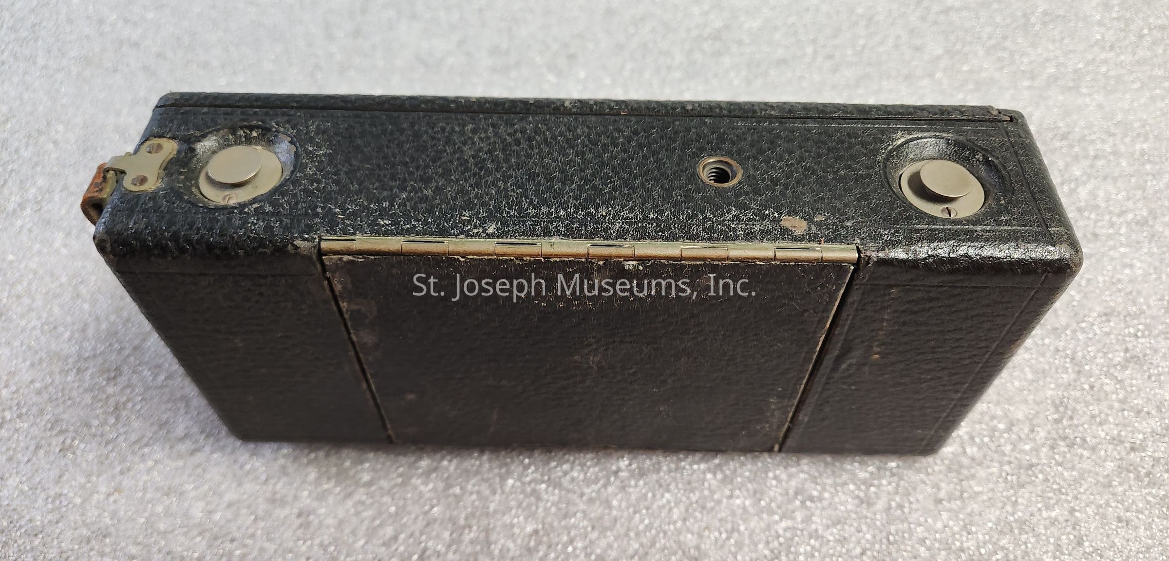 The image shows a vintage black box camera with a textured surface, characterized by worn edges possibly from age or use. On the top side, two circular metallic buttons are visible, which are likely controls for operating the camera. A metallic hinge runs horizontally across the center, suggesting that the camera may fold or open for film loading or viewfinder access. The worn finish and subtle rust on the hinge evoke a sense of nostalgia, hinting at the device's historical or collectible significance, possibly dating back to an era when film cameras were prevalent, marking it as an artifact of photographic history.