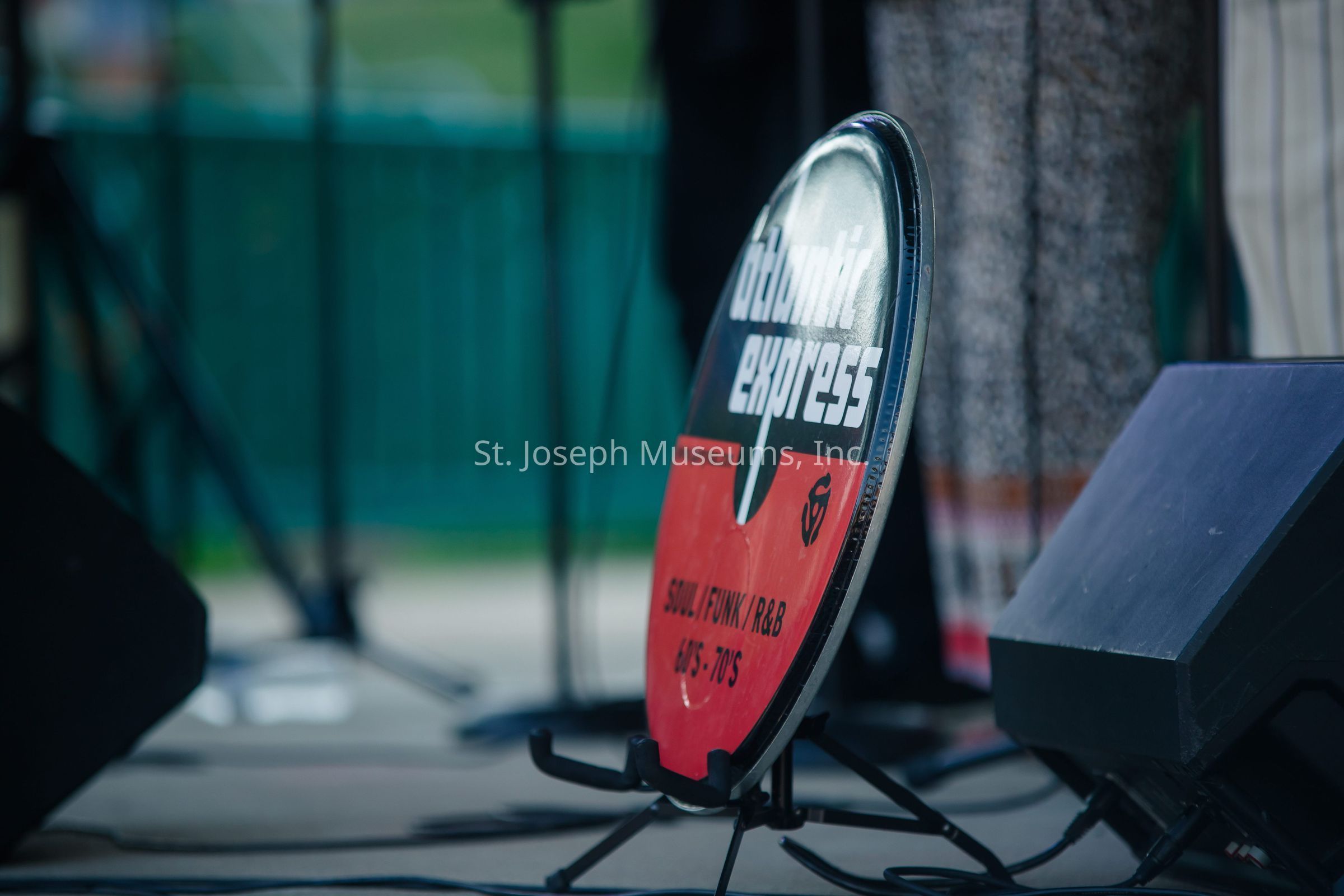 The image captures a close-up of a circular sign positioned on a stand, prominently displaying the text "Galactic Express" in bold, stylized lettering at the center. The sign, which appears to be part of a stage setup, has a retro design with a red and black color scheme, invoking a vintage feel suitable for a music band or performance group. Beneath the main text, additional information reads "Soul / Funk / R&B 60's - 70's," indicating the musical genres and era the group might be associated with. The background of the photograph is out of focus, showing blurred details of a stage or outdoor setting, suggesting that the sign is placed in a live performance environment. Nearby, parts of equipment and instruments such as microphones and speakers are visible, further hinting at a musical context. The overall composition and the elements within the frame create an atmosphere of anticipation for a live musical experience, celebrating classic soul and funk rhythms.