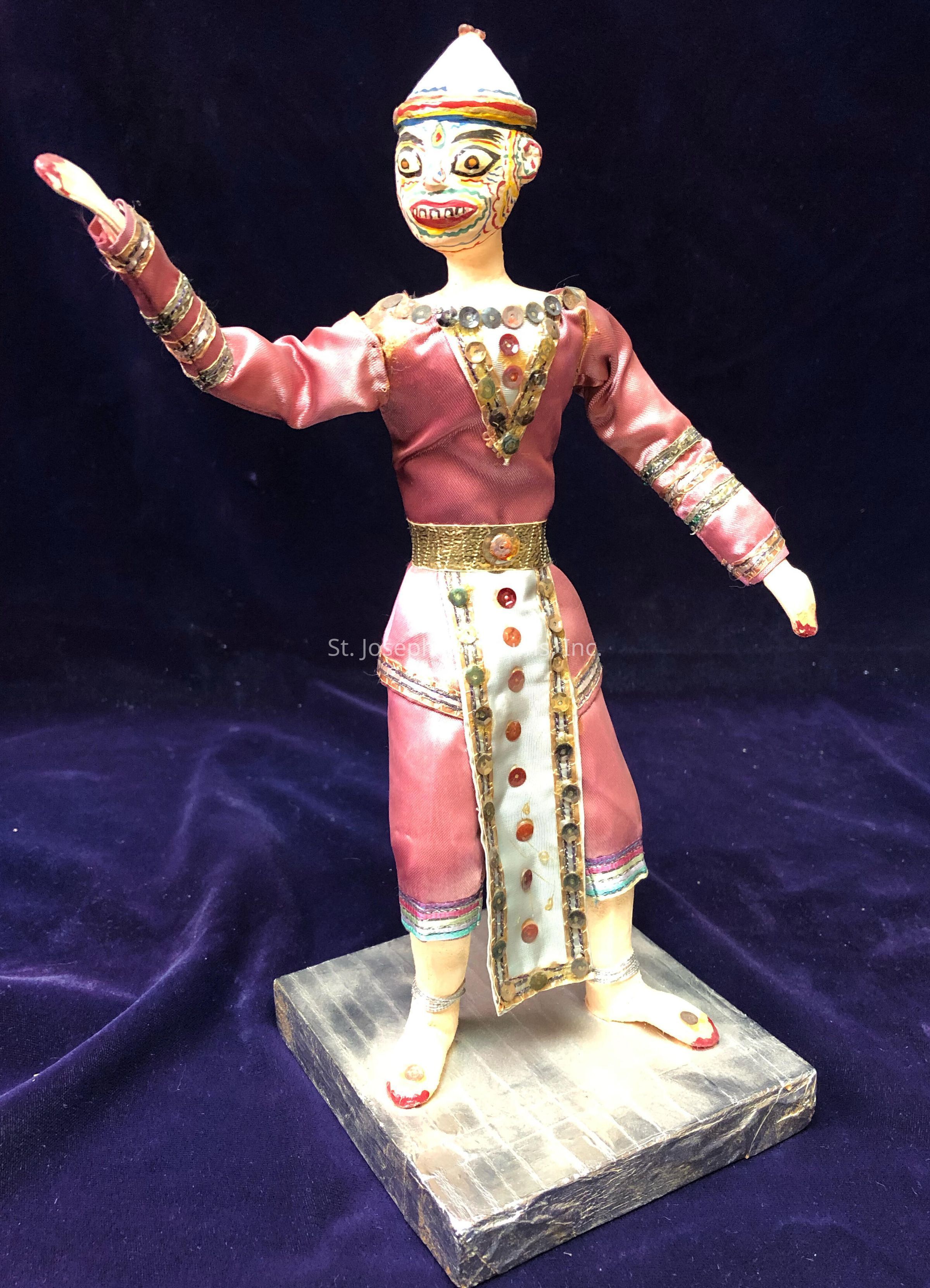The image depicts a colorful figurine dressed in a vibrant outfit, featuring a combination of materials like fabric and adorned with bright, glossy details. The figure has an expressive face painted with intricate designs and is wearing an elaborately decorated costume in shades of pink, gold, and cream. The attire includes a long robe embellished with shiny buttons and intricate trims, accentuating its attention to detail. The figure stands on a square pedestal, and its pose gives the impression of movement or storytelling, with one arm extended outward. The background is a deep, plush purple, which contrasts elegantly with the colorful dress of the figurine, highlighting its craftsmanship and cultural significance.