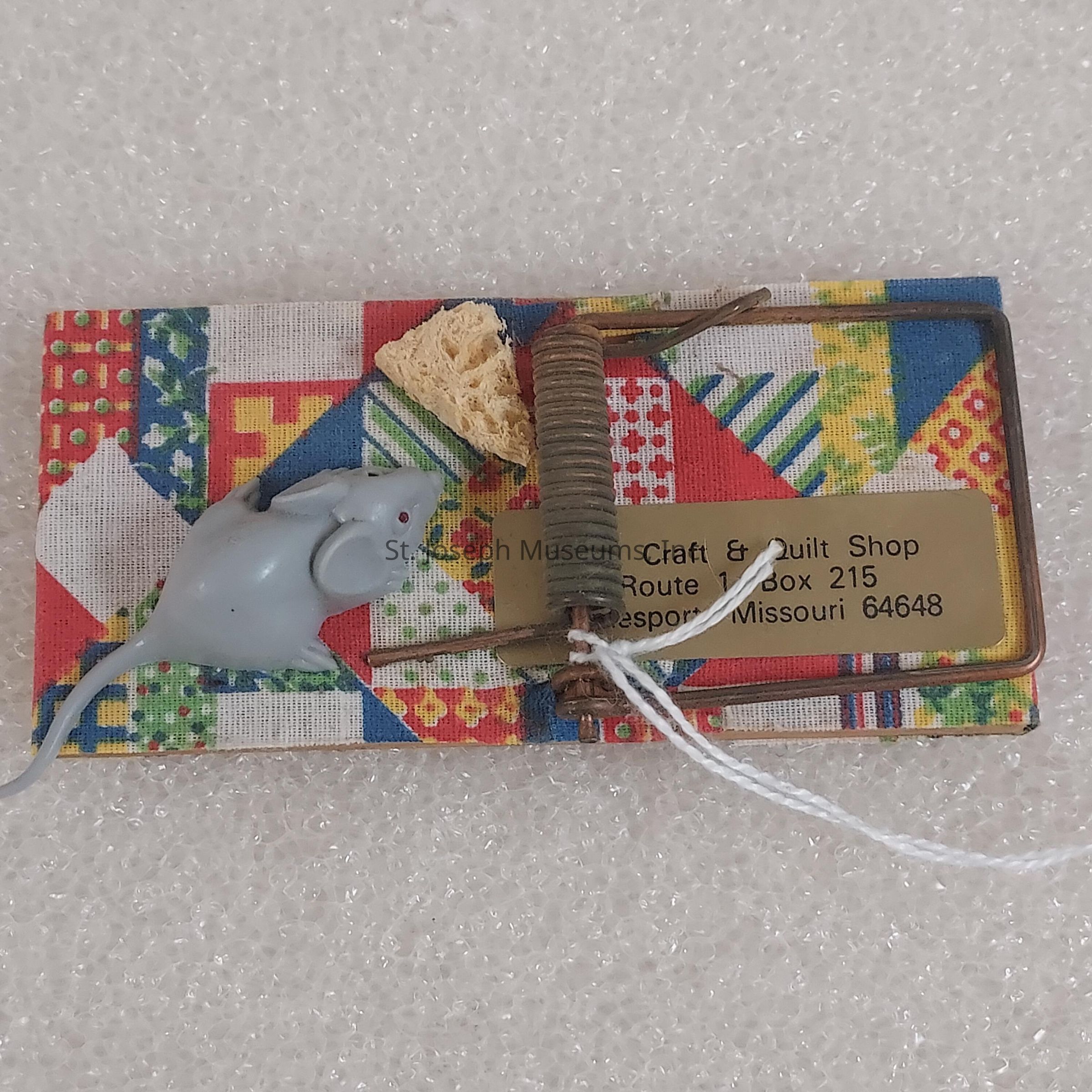 The image depicts a novelty item resembling a decorated mousetrap with a patchwork quilt-like design base, featuring various colorful patterns including stripes and geometric shapes. A small, toy mouse made of plastic is attached to the trap, seemingly as a humorous element. There is a piece of sponge-like material placed on the trap's trigger, mimicking bait. Additionally, the trap includes a metallic plaque with engraved text that reads "Craft & Quilt Shop Route 1, Box 215, Jamesport, Missouri 64648," suggesting it might be a promotional item or souvenir from this location. The overall item rests on a textured, white surface.