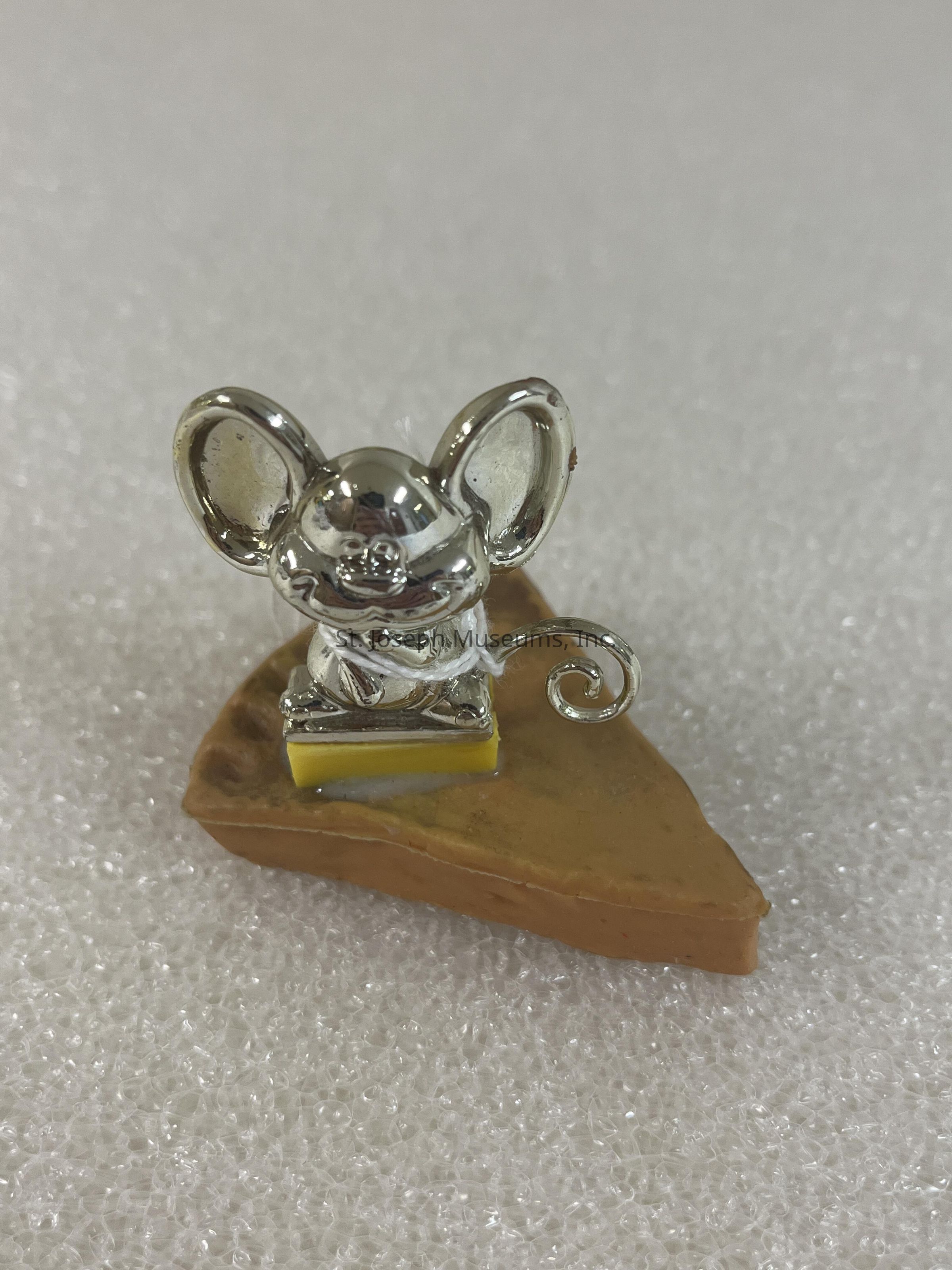 The image depicts a small metallic mouse figurine with large, shiny ears and a gleaming body, perched atop a triangular piece resembling a wedge of cheese. The mouse's body is crafted with intricate details, including a coiled tail, and it sits on a small platform lined with a yellow strip, enhancing its visual appeal. This whimsical figurine is placed on a textured white surface that adds contrast and highlights its polished silver color, creating a playful and charming composition that captures attention with its creative blend of materials and imaginative design.