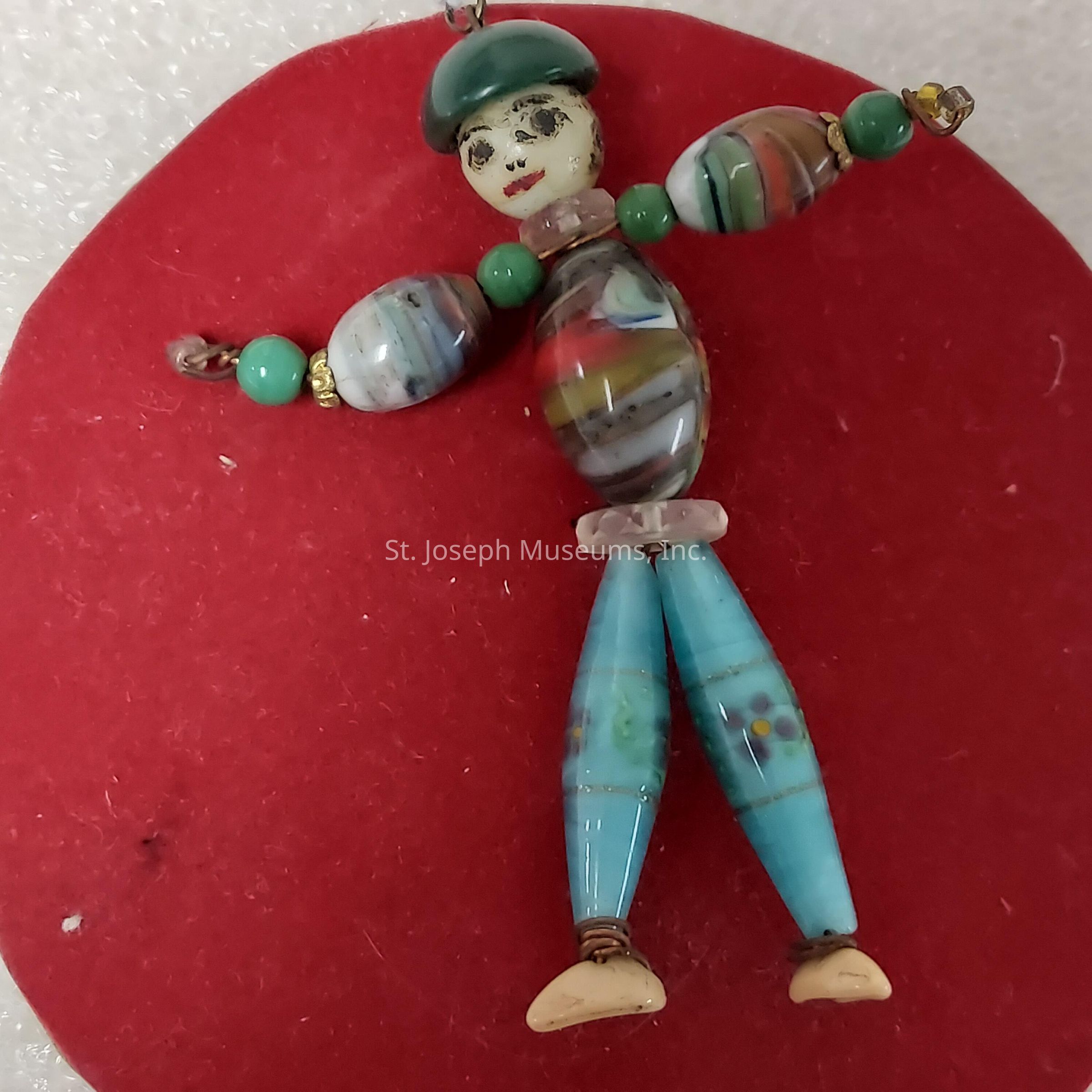 The image shows a whimsical figurine crafted from a variety of colorful beads arranged to resemble a person, with the body made from an oval bead featuring a swirl of multicolored patterns, and arms and legs comprising elongated beads in shades of turquoise and multicolored designs; the head is represented by a small, round bead with painted facial features depicting eyes and a mouth, topped with a green cap bead, creating an overall playful and artistic appearance, all placed against a bright red circular background that enhances the vivid colors of the figurine, offering a striking contrast that serves to highlight the intricate beadwork and playful character of this unique handcrafted item, which also suggests potential as a decorative accessory or artistic piece, and evokes a sense of creativity and charm, making it an engaging and eye-catching visual composition.