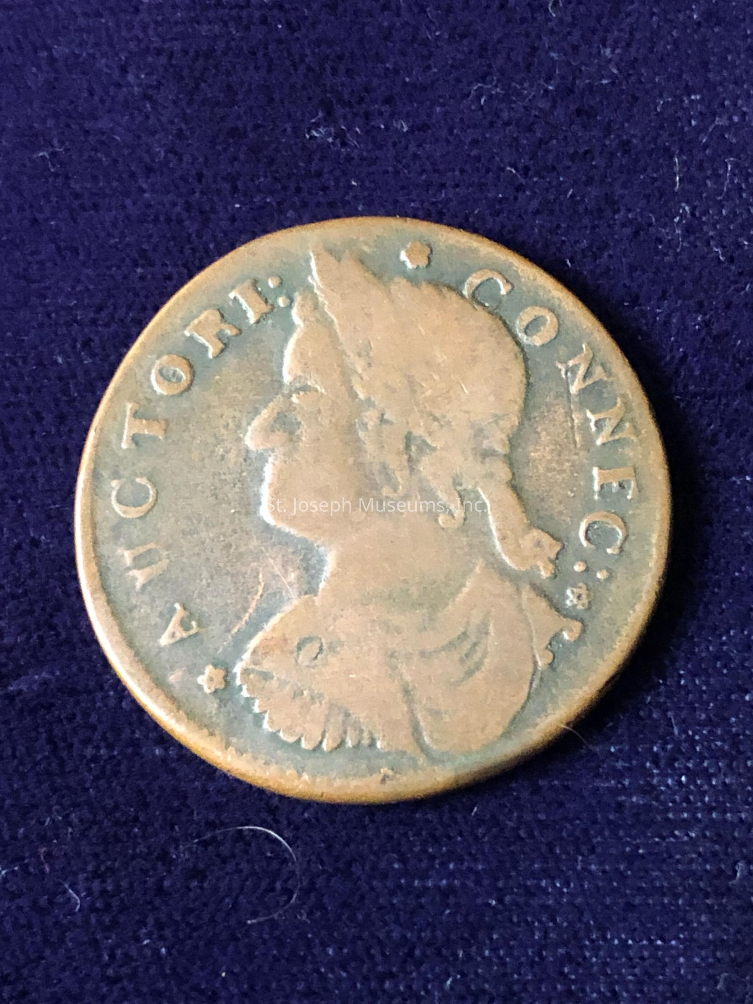 The image displays an antique coin with a worn, copper-like patina, depicting the profile of a figure facing left. Encircling the profile is the inscription “AUCTORI: CONNEC:”, which suggests historical significance, possibly indicating it is a colonial coin from Connecticut. The surface of the coin shows some signs of age and wear, with small imperfections and a slightly textured appearance that gives it a sense of historical authenticity. This artifact may have been from a period where such coins were common in trade, reflecting the era's design styles and minting techniques. The coin rests on a dark blue fabric that contrasts with its metal tone, highlighting its details and contours effectively. A few stray fibers are visible on the fabric, adding a sense of realism to the context in which the coin is placed. It serves as a physical reminder of past economies and trades, inviting curiosity about the stories and transactions it may have been part of during its time in circulation.