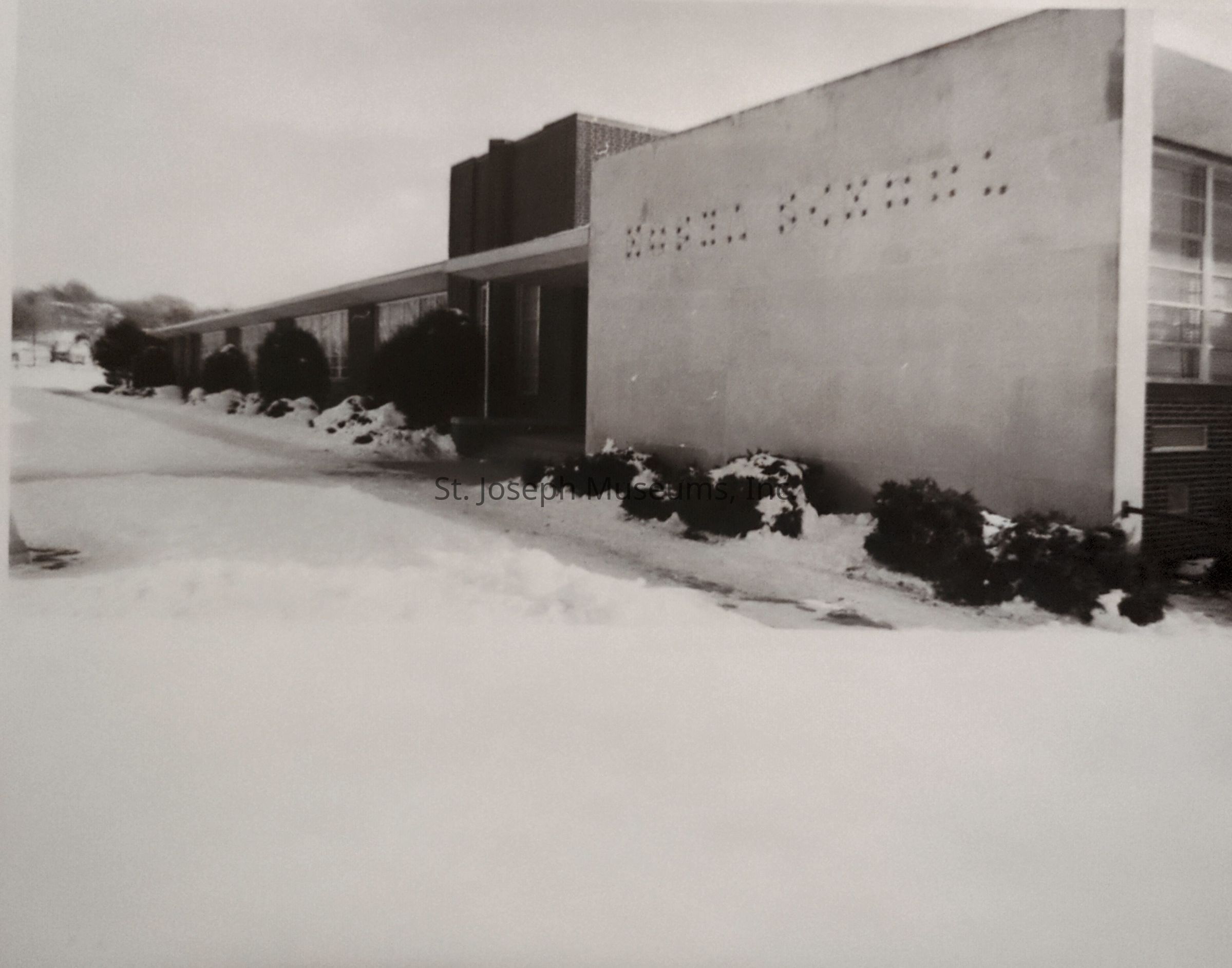 Black-and-white image depicting a modernist building at the corner of Hyde Park and Gordon Avenue in St. Joseph, Missouri. The structure is surrounded by snow-covered ground and shrubs, creating a muted, serene winter scene. The building features large windows and a flat roof, embodying mid-century architectural elements.