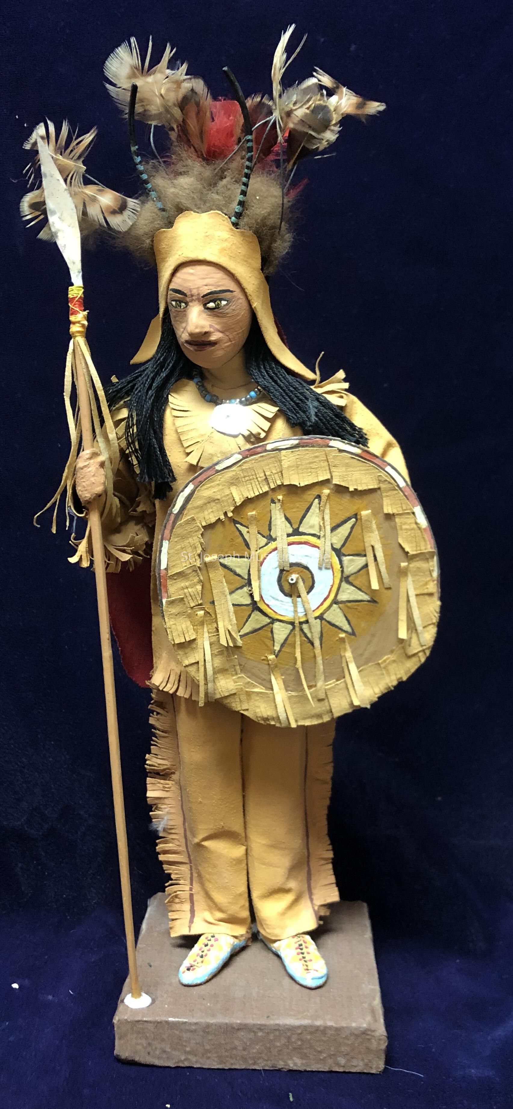 The image shows a handcrafted figurine of a person dressed in traditional attire. The figure is wearing a tan outfit with intricate fringe details and holds a spear in one hand, adorned with feathers. The figure's head is topped with a headdress made up of multicolored feathers and what appear to be horns. In the other hand, the figure carries a shield featuring a vivid sunburst pattern with concentric circles in the center, decorated with fringed elements. The figurine's hair is long and black, and it appears to be wearing a beaded necklace. The base on which the figure stands is a simple brown block, contrasting with the deep blue background. The vibrant details and textured materials suggest an emphasis on traditional craftsmanship, capturing a sense of cultural and historical significance. The entire appearance of the figurine reflects careful attention to detail, from the clothing patterns to the facial expressions painted onto the face, conveying a sense of identity and cultural pride.