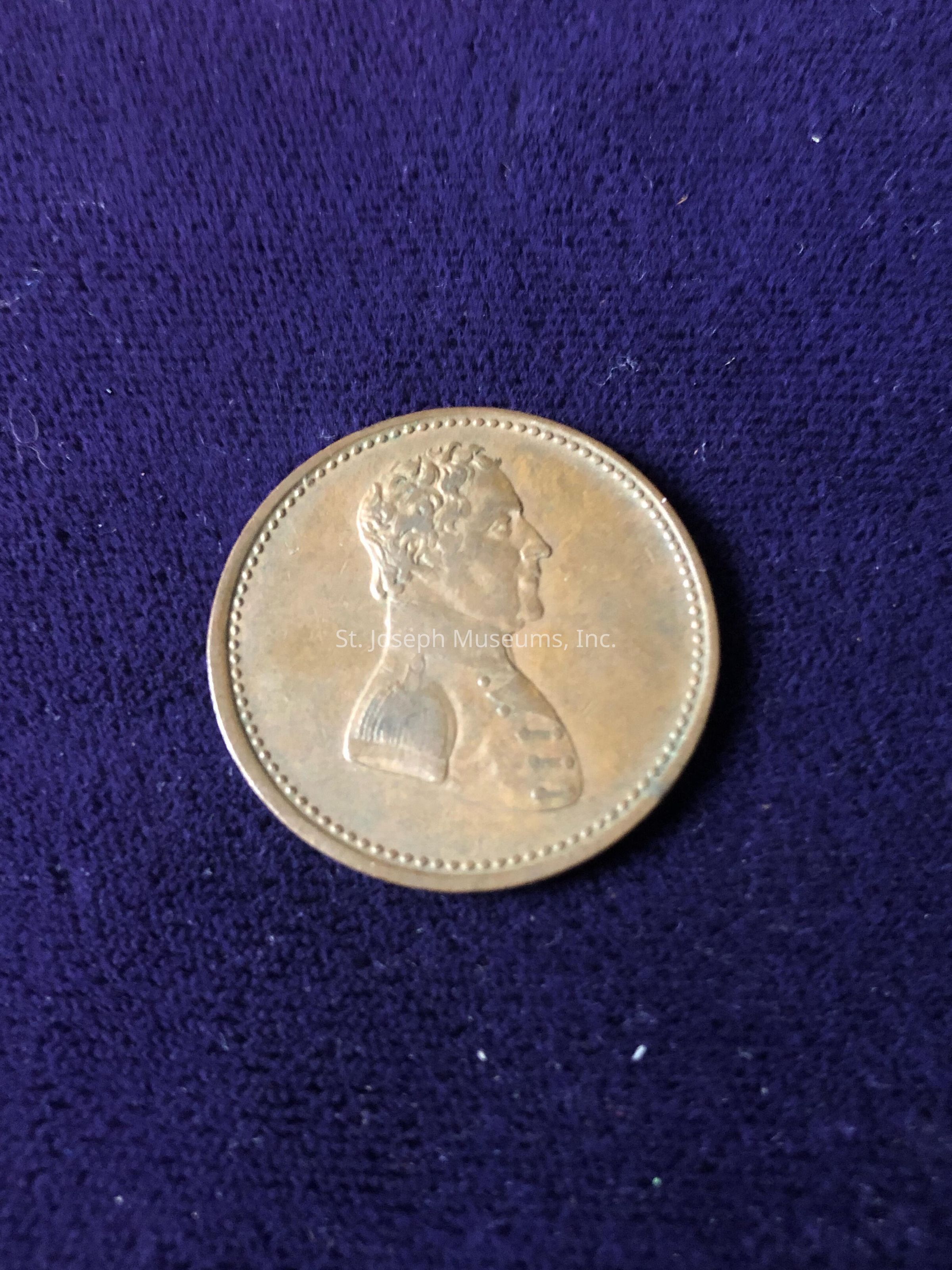 This profile is detailed with short, curly hair, and the attire suggests formal or historical significance, possibly indicating that the coin commemorates a notable figure or event. The coin's design is bordered by a dotted line around its edge, giving it a classical appearance. The warm, metallic hue of the coin contrasts with the rich, dark fabric beneath, highlighting the coin's intricate details and craftsmanship. The fabric surface creates a soft, elegant backdrop that accentuates the coin's distinct features. The lighting in the image gently illuminates the coin, casting subtle shadows that enhance the three-dimensional aspect of the raised profile on the surface. Overall, the composition captures the essence and historical aura of the coin, making it a focal point against the luxurious fabric background, which highlights its possible numismatic value.