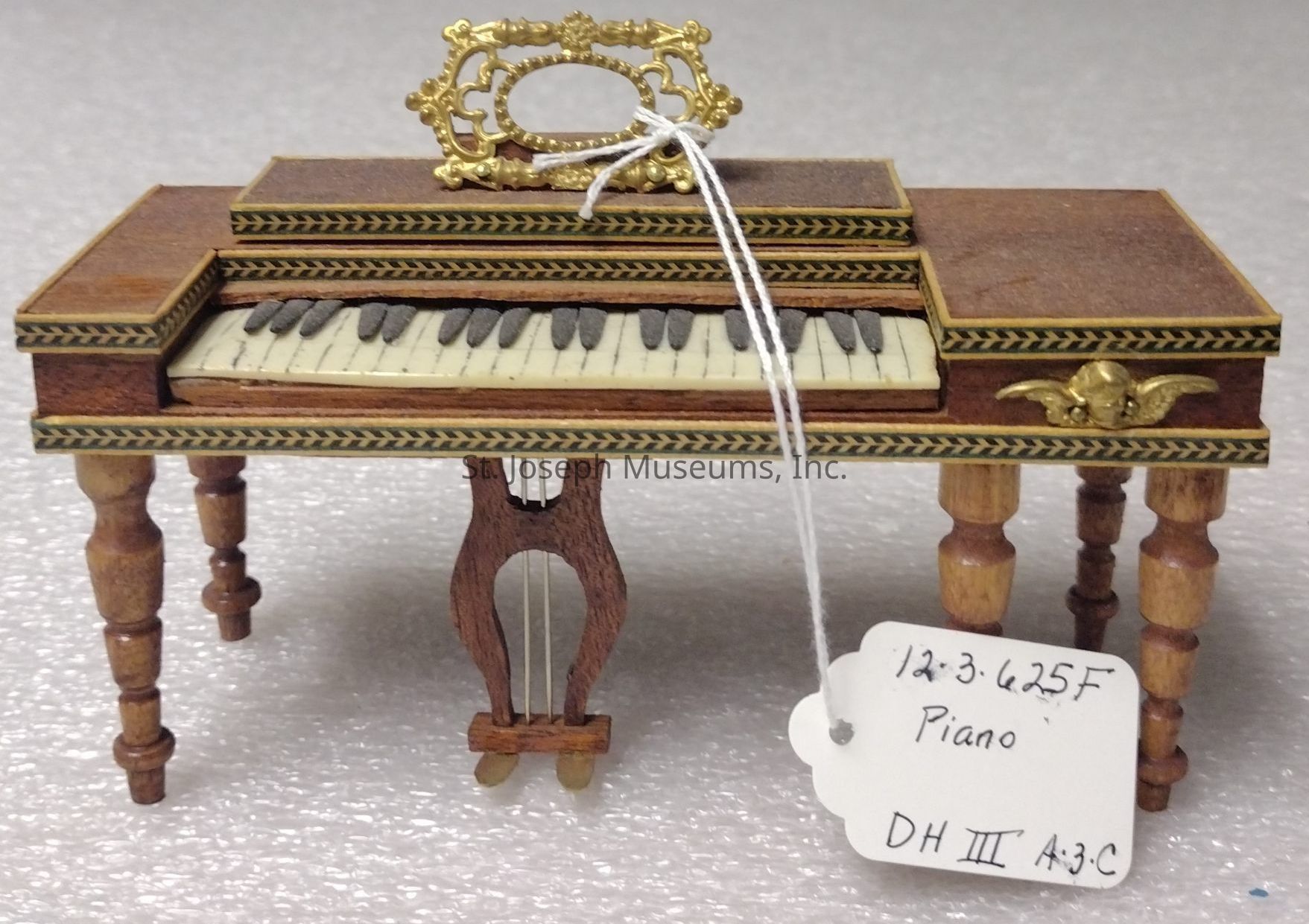 The image depicts a miniature piano crafted with intricate wooden details, featuring an ornate gold-colored music stand and adorned with a decorative pattern along its edges. The piano has four delicately turned wooden legs, adding to its vintage charm. Resting on top is an elegant music sheet holder crafted with golden designs. A small white string is attached to the piano's upper structure, holding a tag labeled "12-3-625F, Piano, DH III A3-C." The miniature has a realistic representation of the white and black keys, complemented by a textured background, enhancing its authenticity as a scaled-down musical instrument.