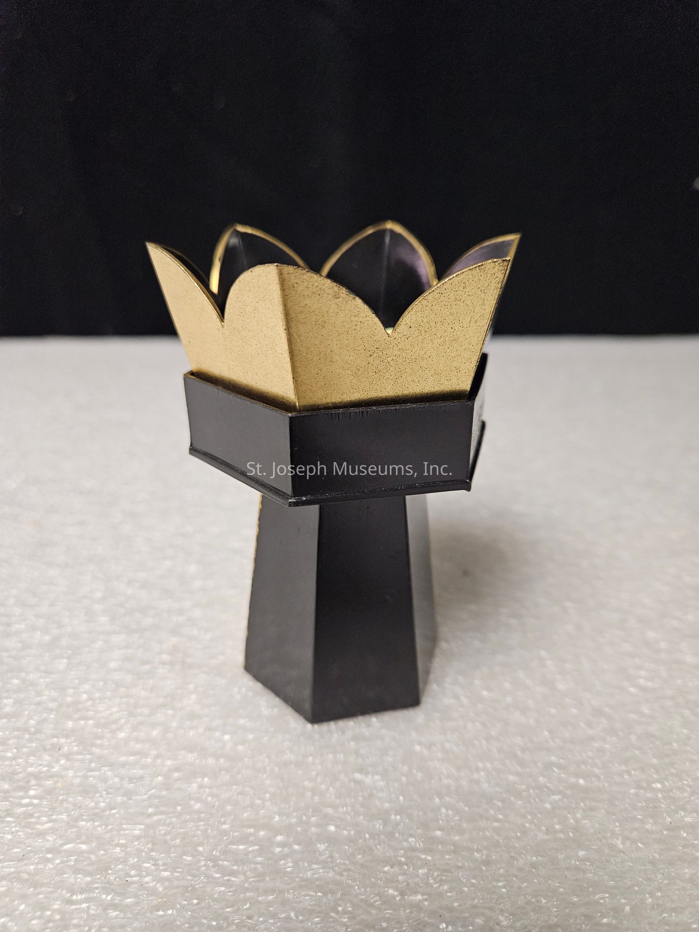 The image depicts a small, artistic object resembling a cup or pedestal with a geometric and modern design, placed against a contrasting background of dark and light tones. The structure features a black base with a tall, narrow shape tapering towards the top, which transitions into a hexagonal platform supporting golden, petal-like extensions that appear both metallic and glossy. These ascending petals create a visually striking crown-like appearance, harmonizing with the sleek sharpness of the overall form. The object rests on a textured, white surface that appears soft or foam-like, providing a balanced contrast to the stark, black vertical element. Meanwhile, the backdrop is a smooth, deep black, effectively drawing attention to the item’s vivid elegance and intricate detailing. The composition and lighting focus on accentuating the harmonious blend of matte and glossy textures, highlighting the mesmerizing simplicity and elegance of the object’s unique design.