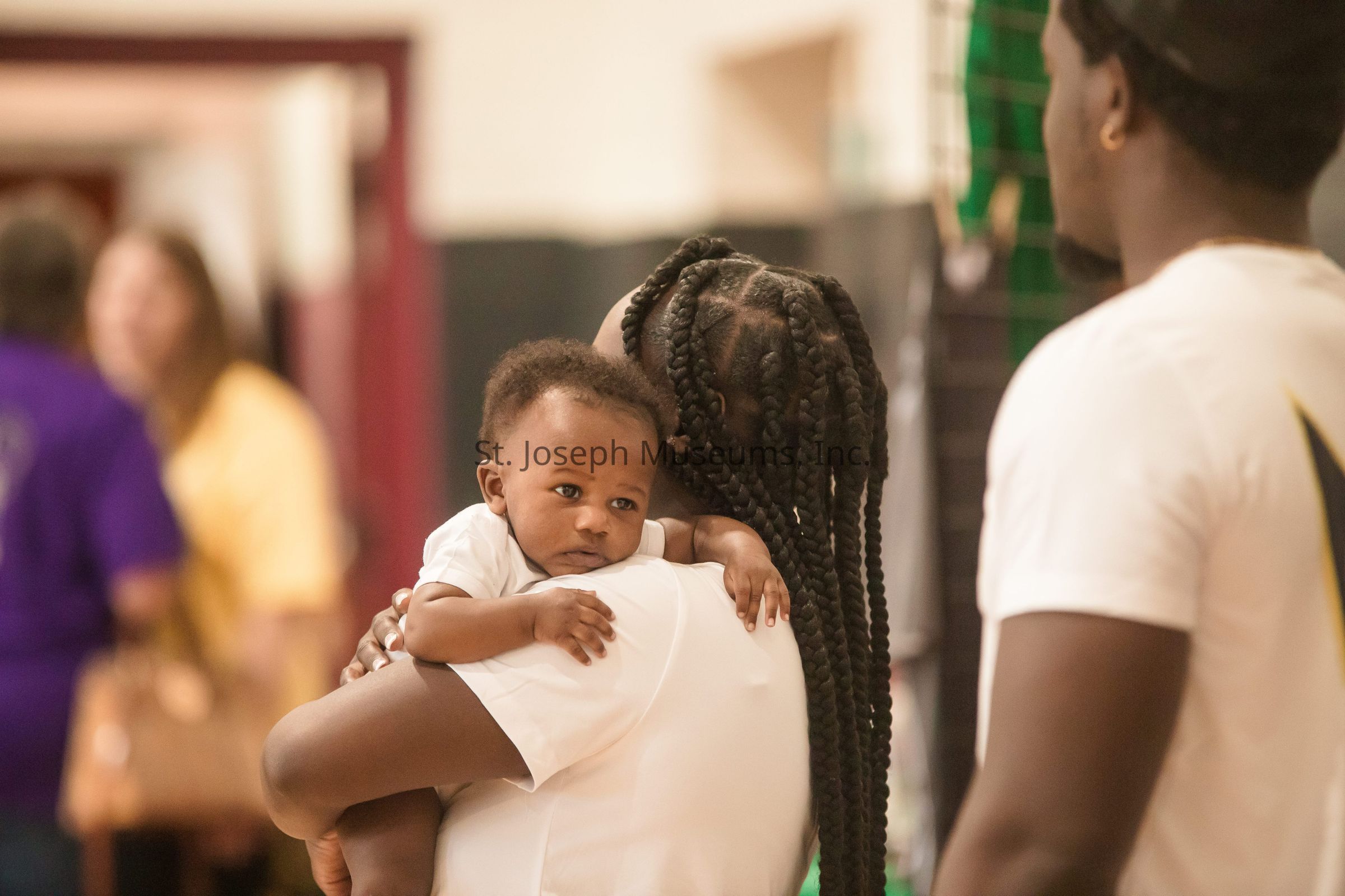 In the image, a woman with beautifully braided hair is tenderly holding a baby on her shoulder while standing in what appears to be an indoor setting; the baby, wearing a white outfit, has a serene expression and rests comfortably against the woman, who is wearing a white shirt, as another person in the foreground, also dressed in white, stands nearby facing them; the background shows blurred figures and various colors, suggesting a lively environment, possibly a community gathering or event, where people are engaged in conversations and activities, creating a warm and familial atmosphere.