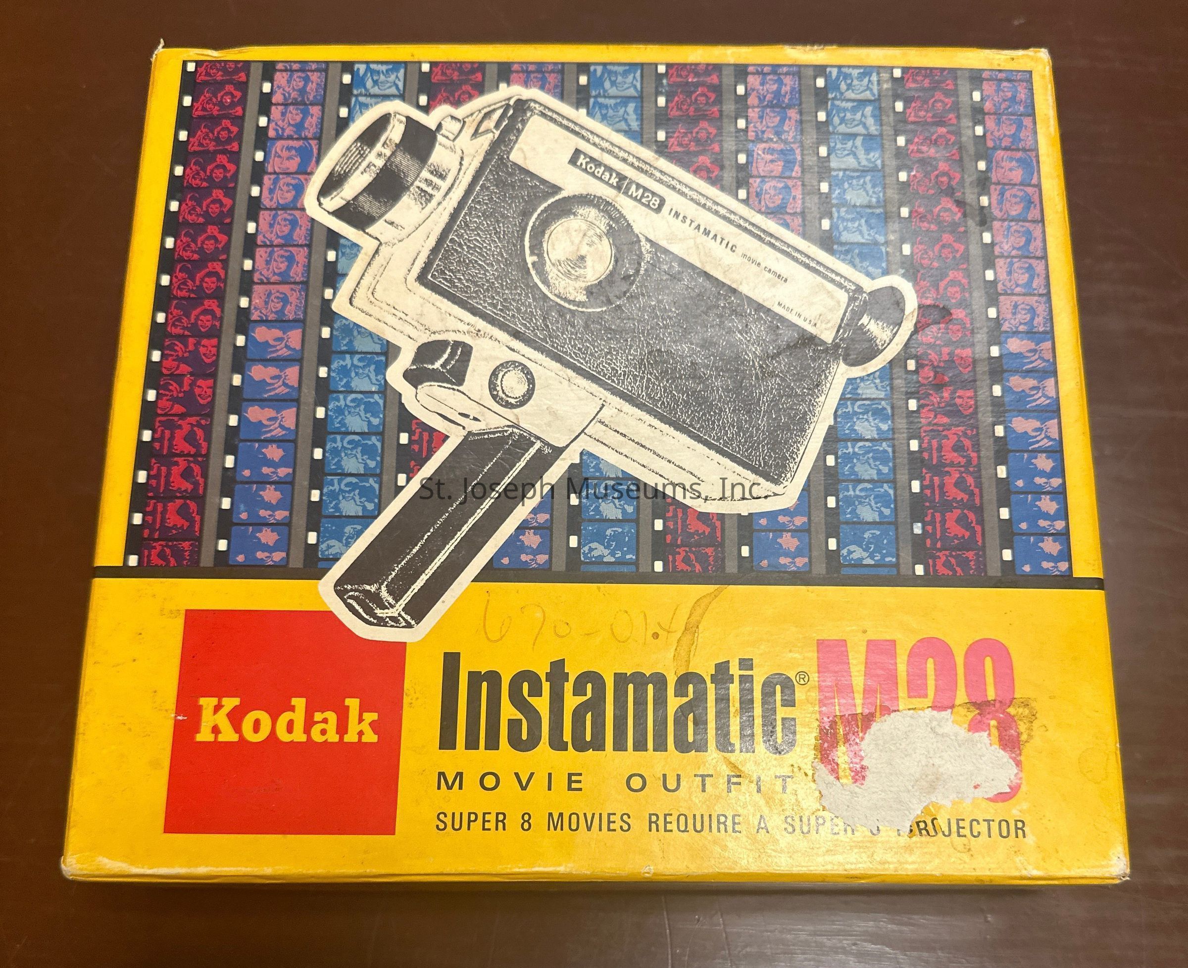 A: The image showcases a vintage Kodak Instamatic M28 movie camera box, featuring a prominent illustration of the camera in a black and white sketch style. The box has a vibrant yellow background, accented by a decorative strip at the top with red and blue film reel patterns. The Kodak logo appears in a bold red square on the left, while the words “Instamatic M28 Movie Outfit” are printed prominently in black and pink on the right side. The text indicates that Super 8 movies require a Super 8 projector, emphasizing the product's linkage to the Super 8 film format, which was popular during the mid-20th century.