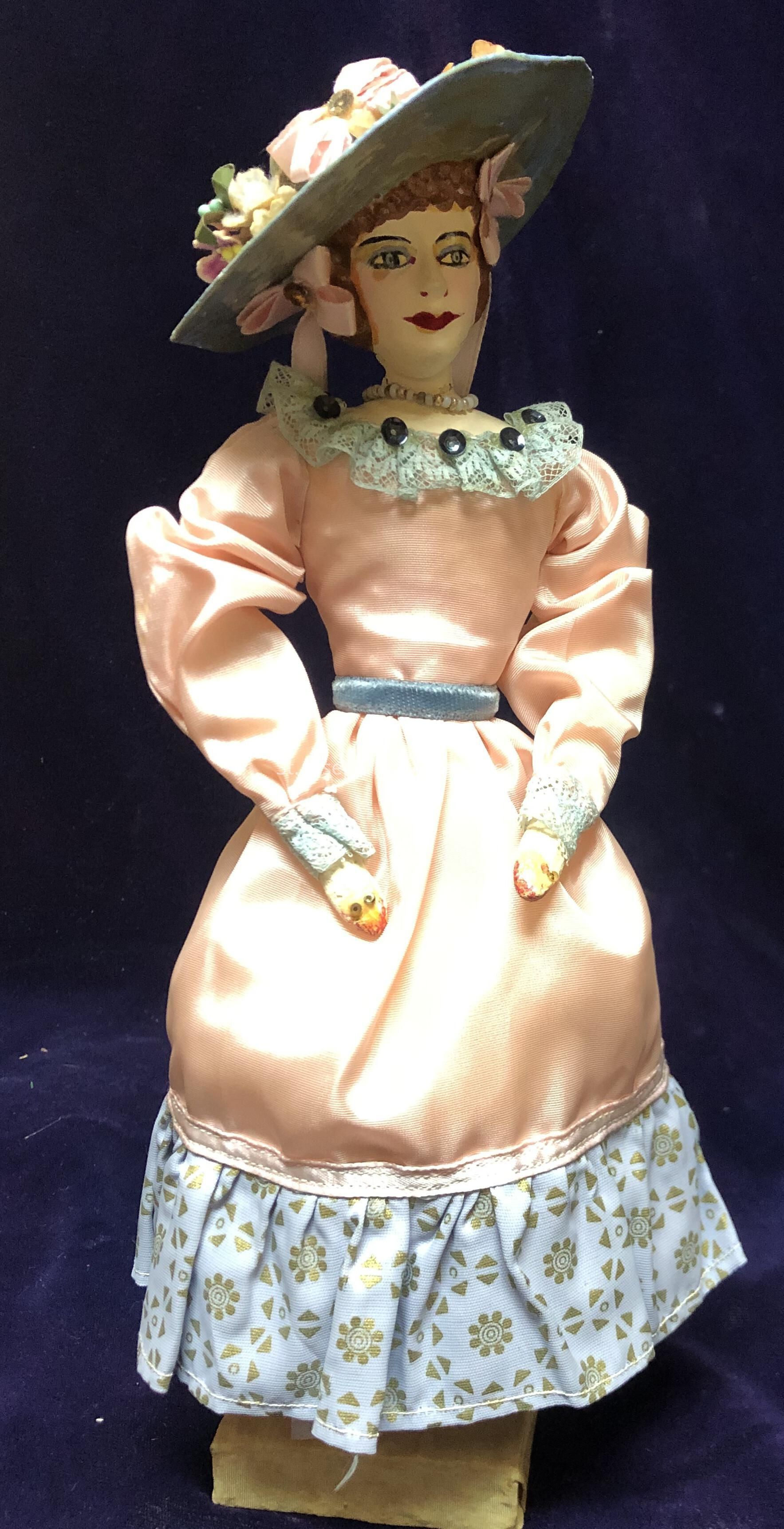 The image depicts a vintage doll wearing an elaborate, elegant outfit. The doll is adorned with a wide-brimmed, flower-adorned hat that adds to the sophistication of the ensemble. The doll's dress is a combination of peach satin and patterned fabric, featuring puffed sleeves and a fitted waist accentuated by a blue belt, enhancing its Victorian-inspired design. Lace detailing decorates the collar and cuffs, adding a touch of delicacy. The doll has a painted face with bold makeup, including red lips and defined eyebrows, emphasizing a classic, graceful look. Standing against a dark backdrop, the doll exudes an air of timeless elegance.