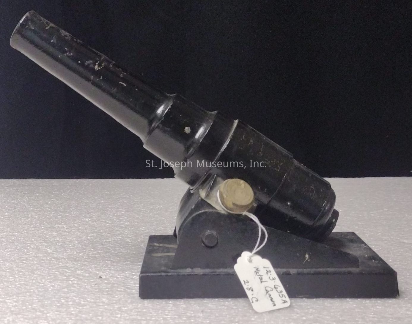 The image shows a small black object resembling a miniature cannon or signal gun mounted on a rectangular base. The body of the object appears cylindrical with a slightly tapered barrel extending outward, suggesting it might be an antique model or replica. Its surface is worn, indicating age or frequent handling, and it is affixed with a rounded brass fastener near its midpoint for support or adjustment. Attached to the base is a small white paper tag marked with handwritten text, which may denote its catalog information or identification number, suggesting it could be part of a collection, perhaps within a museum or exhibit setting. The textured surface beneath gives the impression of a display setup, likely intended to highlight the object against a plain, softly illuminated backdrop, enhancing its visual emphasis. Overall, the combination of materials and the slightly matte finish contribute to a vintage aesthetic, hinting at its historical or ornamental value, while the stark contrast between the black object and the lighter surroundings draws immediate attention to its distinctive structure and craftsmanship.