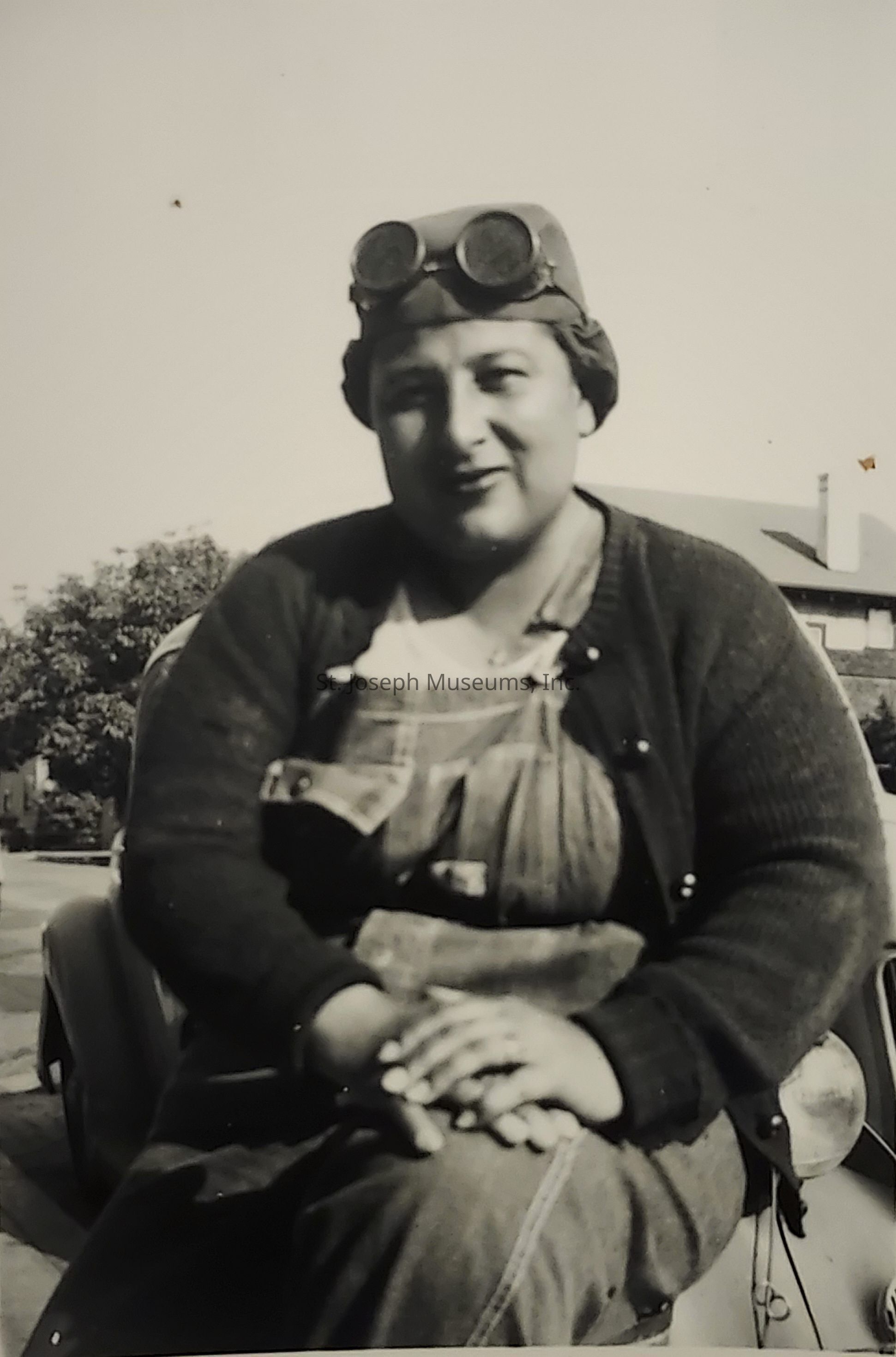The black-and-white image depicts a person sitting on the front of a vintage vehicle, possibly from the early 20th century, as indicated by the style of their attire and accessories. The individual is dressed in work overalls with a cardigan and is wearing a headcover that supports a pair of round goggles, suggesting they might be involved in a mechanical or outdoor activity common in that era, such as driving or maintaining vintage cars. The person sits comfortably with hands folded, expressing a relaxed demeanor. In the background, you can see elements of an urban or suburban setting, with parts of a house and trees visible, hinting at a peaceful residential neighborhood. The photograph captures a moment that reflects the everyday life and fashion of the time, offering a glimpse into the past. The natural lighting and the person's direct gaze give the image a candid and personal feel, evoking curiosity about the individual's story and the context of their environment.