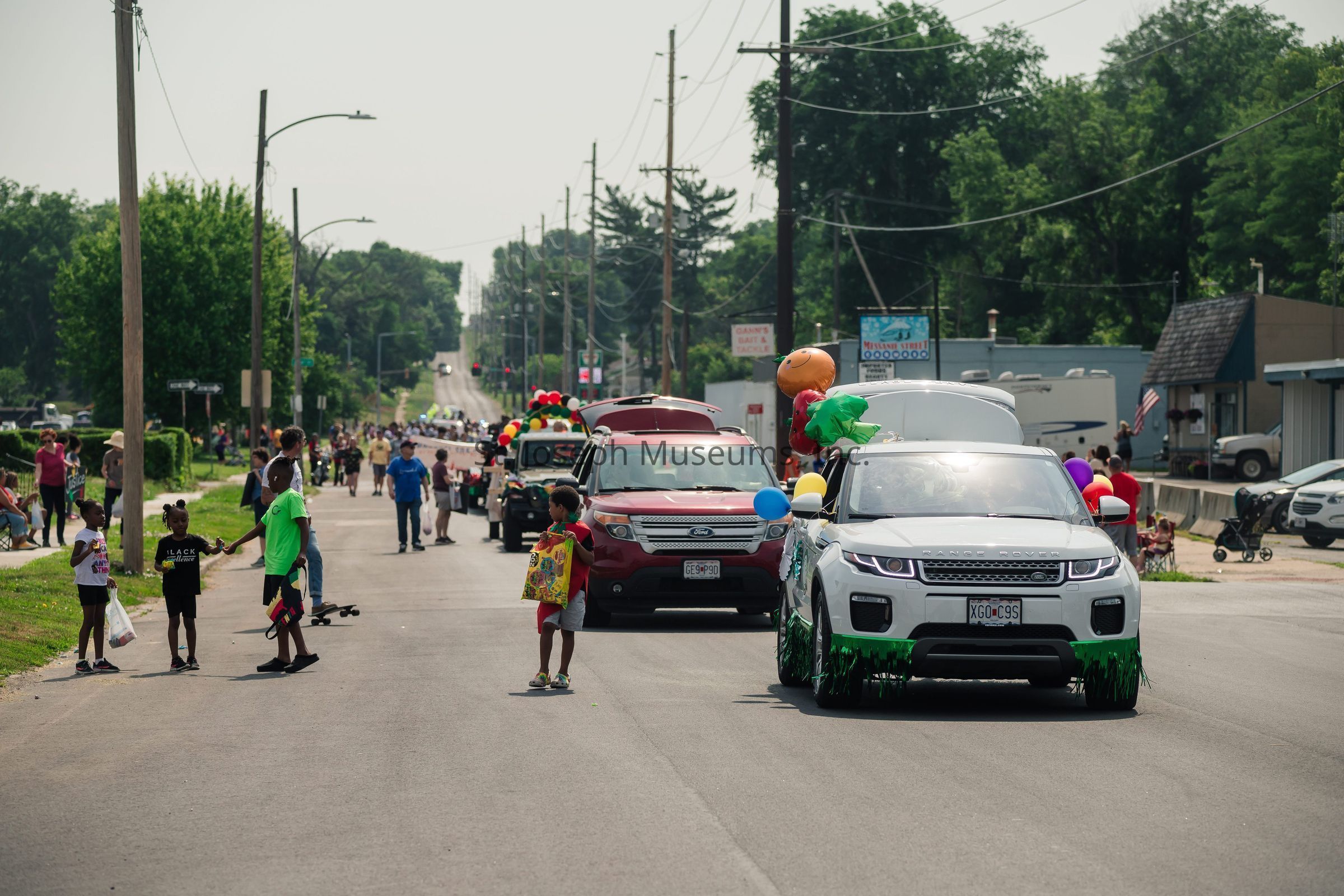 The image depicts a lively parade scene on a wide street, bustling with people and colorful decorations. Various cars, adorned with balloons and festive ornaments, drive along the road. Children and adults are seen walking alongside, some holding bags, likely for catching candy or treats thrown during the parade. In the distance, a group of people walks together, possibly following a banner or float that is part of the parade. The atmosphere is joyous, with a clear sky and abundant greenery lining the street, suggesting a vibrant community event. The whole scene conveys a sense of celebration and togetherness.