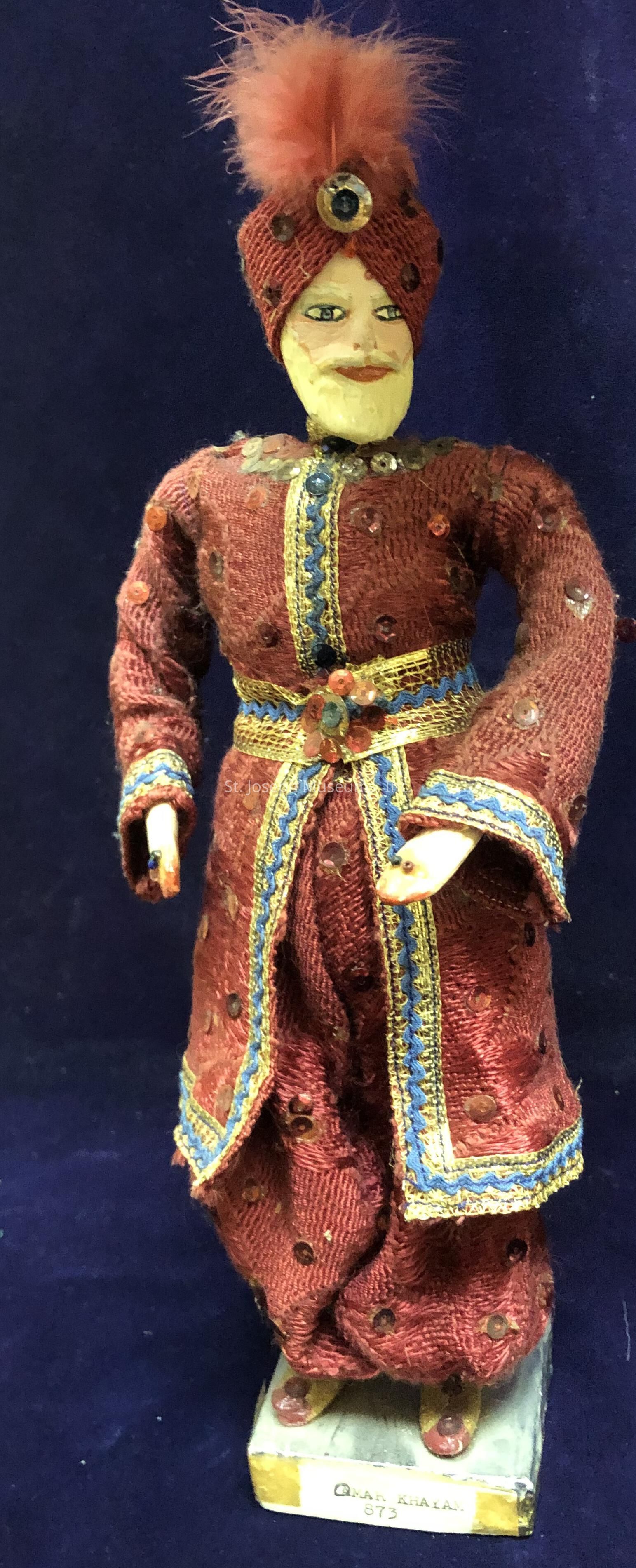 The image shows a detailed figurine dressed in an ornate costume, featuring a maroon outfit embellished with decorative patterns and sequins. The figure wears a matching maroon turban adorned with a large, round jewel and a pink feather plume on top, creating a sense of regality. The edges of the garment are intricately lined with blue and gold trim, contributing to the richness of the attire. A colorful sash with a sparkling floral motif cinches the waist, complementing the outfit's overall elaborate design. The figure, mounted on a rectangular base, stands against a dark, plain backdrop, highlighting its vivid colors and intricate craftsmanship.