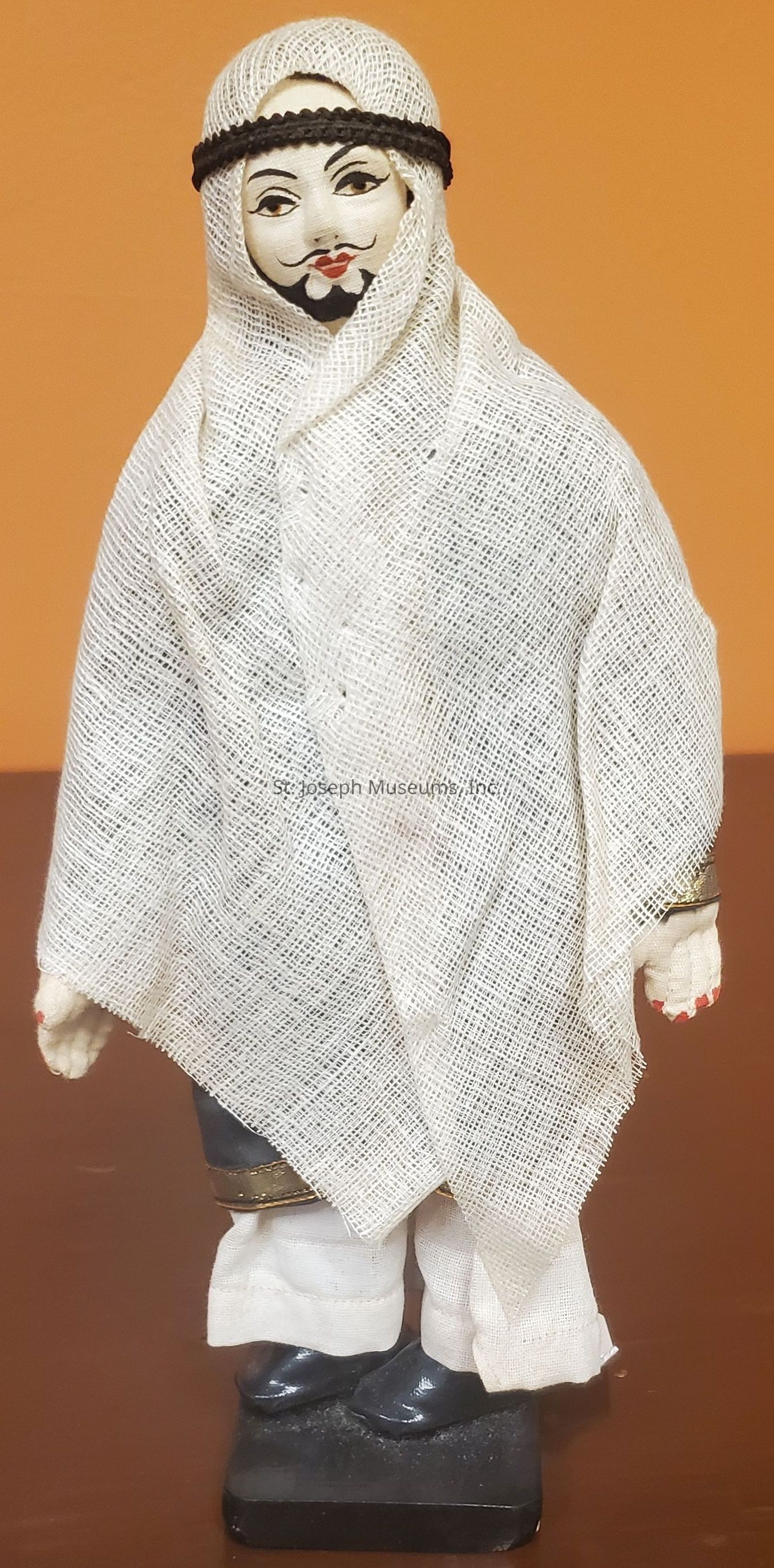 The image shows a figurine dressed in traditional attire, featuring a white, textured fabric draped over its head and body, with only the face exposed. The figure has a painted face with distinctive eyes and a mustache, giving it a culturally specific appearance. The headpiece is secured with a black braided band, and the figurine's lower half displays detailed footwear and layered clothing. The overall craftsmanship suggests a handcrafted quality, emphasizing cultural significance and artistic care, set against a plain orange-brown background, highlighting the figurine’s intricate details and textures.