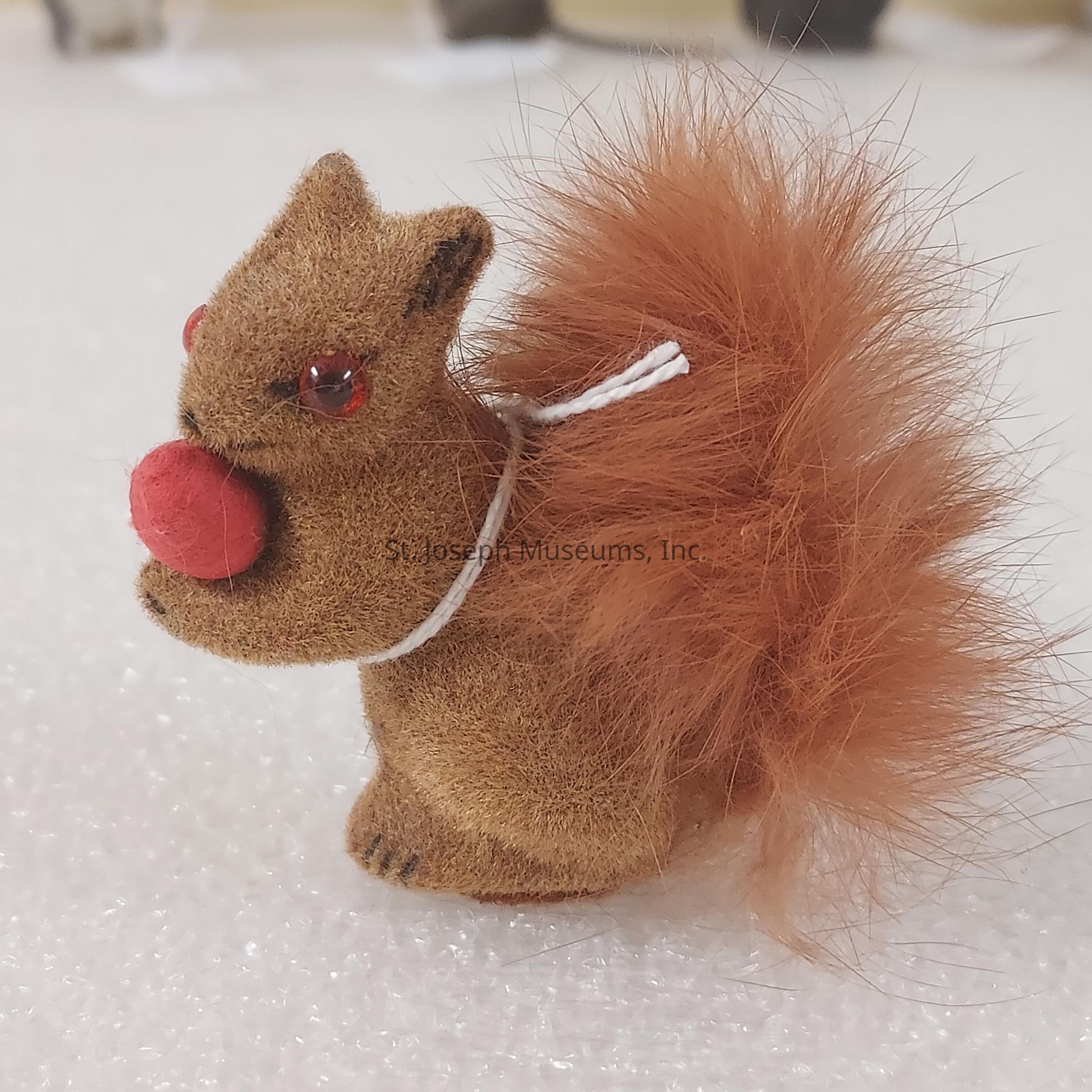 The image showcases a small, fuzzy squirrel figurine sitting on a textured white surface, adorned with a cute red pom-pom nose, shiny red eyes, and a fluffy, bushy tail, which gives it a whimsical and playful appearance enhanced by its soft brown body, slightly rounded ears, and delicate facial details that contribute to its charming, lifelike expression, while a thin white string is looped around its neck, suggesting it might be used as a hanging ornament or decoration; the vivid colors of this delightful object stand out against the plain background, creating a pleasant contrast and drawing attention to its intricately crafted features, which captivate the viewer and highlight the artistry involved in its creation.