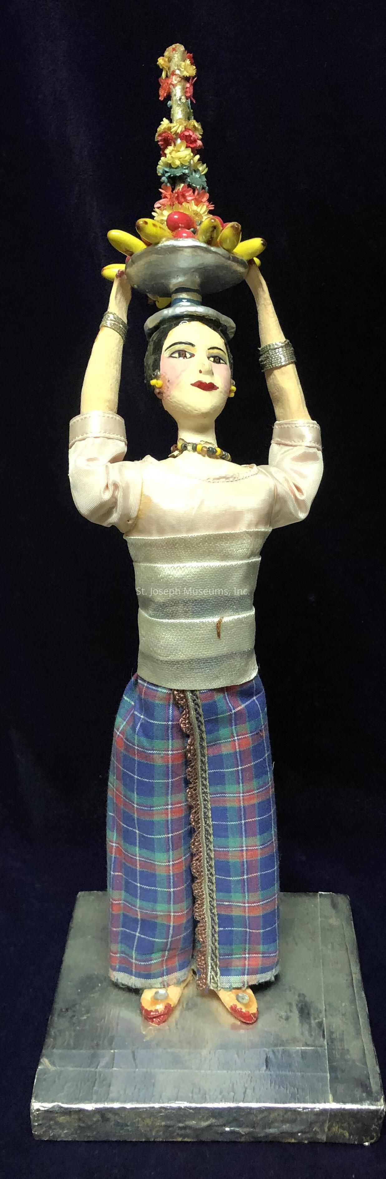 The image depicts a painted figurine of a person standing on a square base, dressed in traditional attire consisting of a white top with long sleeves and a colorful plaid skirt. The figurine is accessorized with a silver belt, matching bracelets, yellow earrings, and a beaded necklace. It is holding a silver tray above its head, filled with a vibrant arrangement of fruits, predominantly yellow bananas, and adorned with multicolored flowers. The tray is gently balanced by both hands, with its decorative handle extending upwards. The outfit and pose suggest a cultural or ceremonial significance, presenting a sense of grace and elegance.