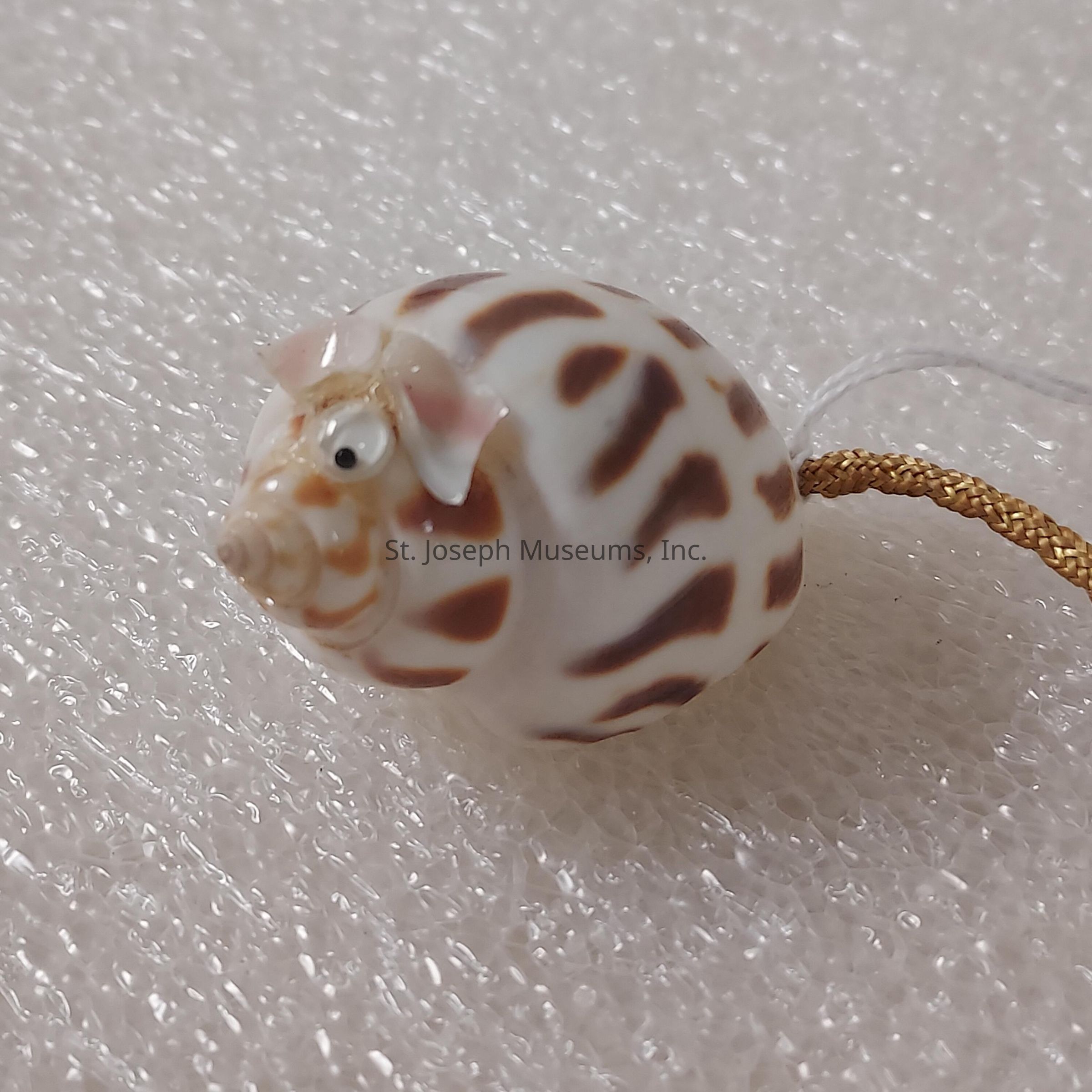 The image showcases a decorative item resembling a pig, crafted from a seashell featuring a glossy surface with white and brown patterns that mimic the natural shell texture. The pig sculpture is detailed with tiny eyes, pink ears, and a small snout, giving it a playful and charming appearance. The shell's inherent spiral and coloration contribute to the design, enhancing the pig's body with a spiraling pattern that adds depth and interest to the piece. It is attached to a thin, golden cord, suggesting it might be used as a hanging ornament or decoration. The surface on which the pig rests appears to be a textured, translucent material reminiscent of bubble wrap or frosted glass, which provides a subtle, soft contrast to the smooth and polished texture of the seashell. The overall composition of the image captures the delightful whimsy of using natural elements to create art, blending intricate handicraft with the inherent beauty of nature.
