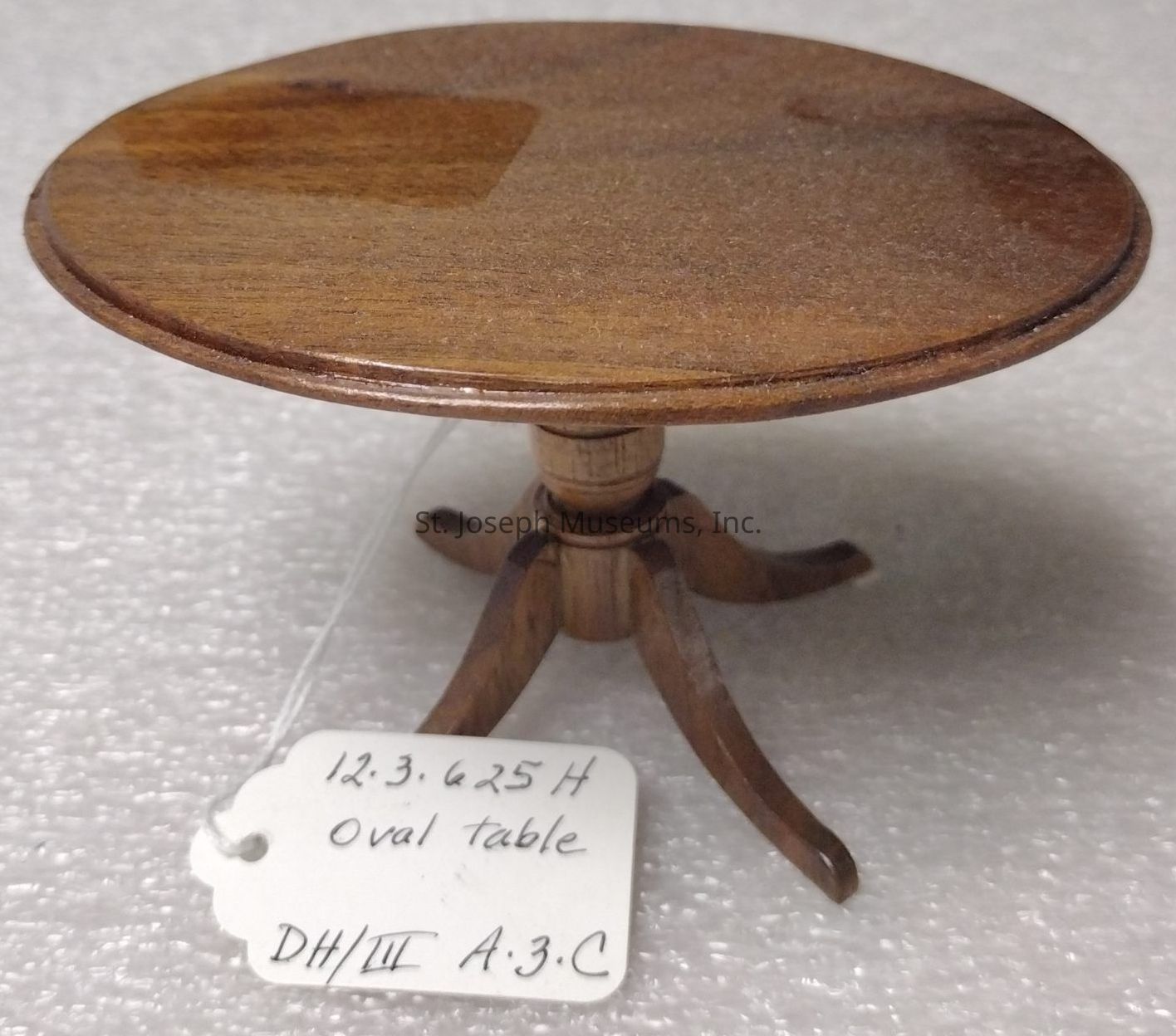 The image showcases a miniature oval table made of wood, featuring a finely polished brown surface that exudes a classic and elegant appearance. The table stands on a central pedestal base comprised of four intricately carved legs, which provide sturdy support while adding to the overall aesthetic appeal. Accompanying the table is a small white tag with handwritten details in black ink, which includes numbers and labels such as "12.3.625 H", "oval table", and "DH/III A.3.C," suggesting identification or cataloging information. The background surface is textured, possibly providing contrast against the smooth finish of the table, highlighting its craftsmanship.