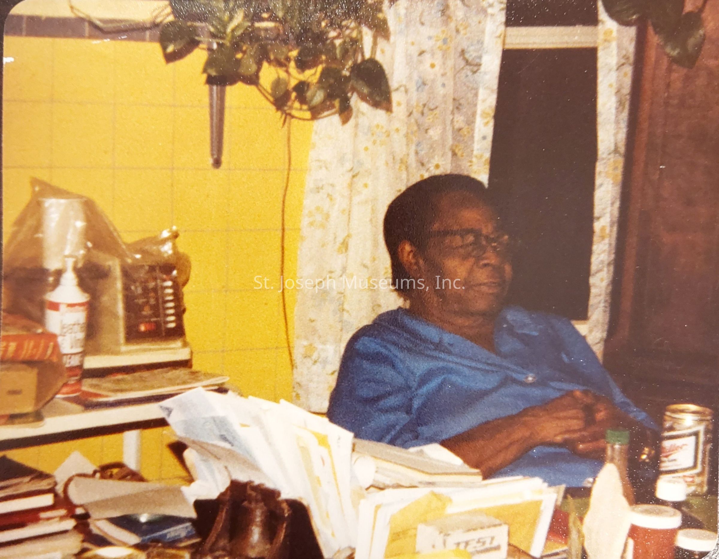 The image depicts a person seated at a cluttered table within a room featuring yellow tiled walls and flowery curtains partially covering a window. The individual, wearing a blue outfit, appears relaxed and is possibly holding or touching something near their hands. On the table, numerous items are scattered, including several stacks of papers and books, an unlabeled can, a bottle of hot sauce, and an old-fashioned radio. A hanging basket, containing a potted plant with trailing vines, is suspended on the wall, adding a touch of greenery to the scene. Additionally, there is a bottle resembling either a cleaning solution or a household product, distinguishable by its red and white label, visible near the radio. This setting, likely a kitchen or a cozy living area, conveys a sense of being lived-in and well-used, capturing a moment of everyday life with an atmosphere of nostalgia, comfort, and personal history.