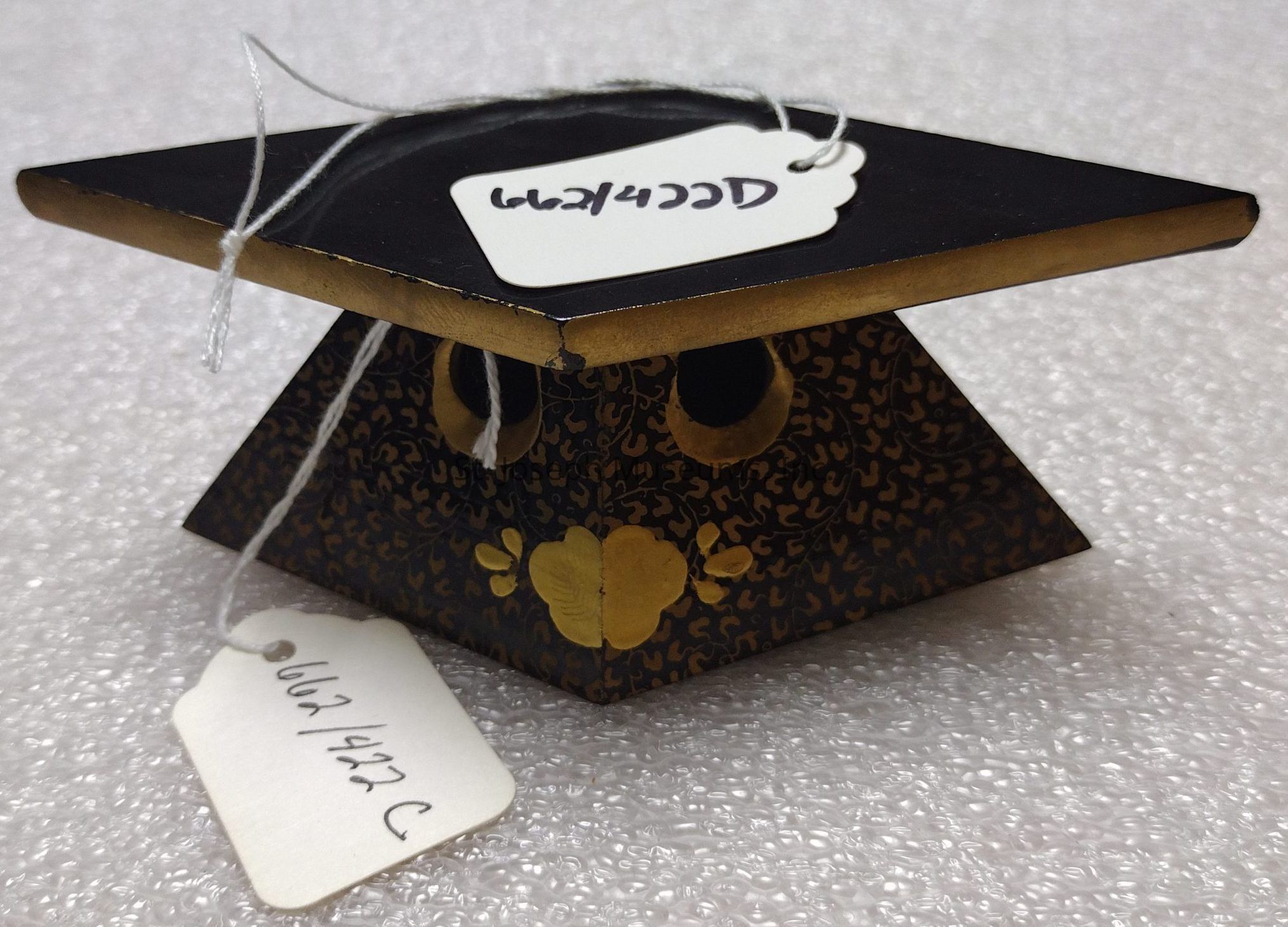 The image features a unique, pyramid-shaped object with a flat, square top, painted in black with gold edges and intricate leafy patterns on its sides. The object is embellished with circular gold accents and a heart-like motif on one side. It is adorned with two white tags connected by strings, each displaying a handwritten code. The object rests on a textured, bubble-wrap-like surface, enhancing its ornate and decorative appearance.