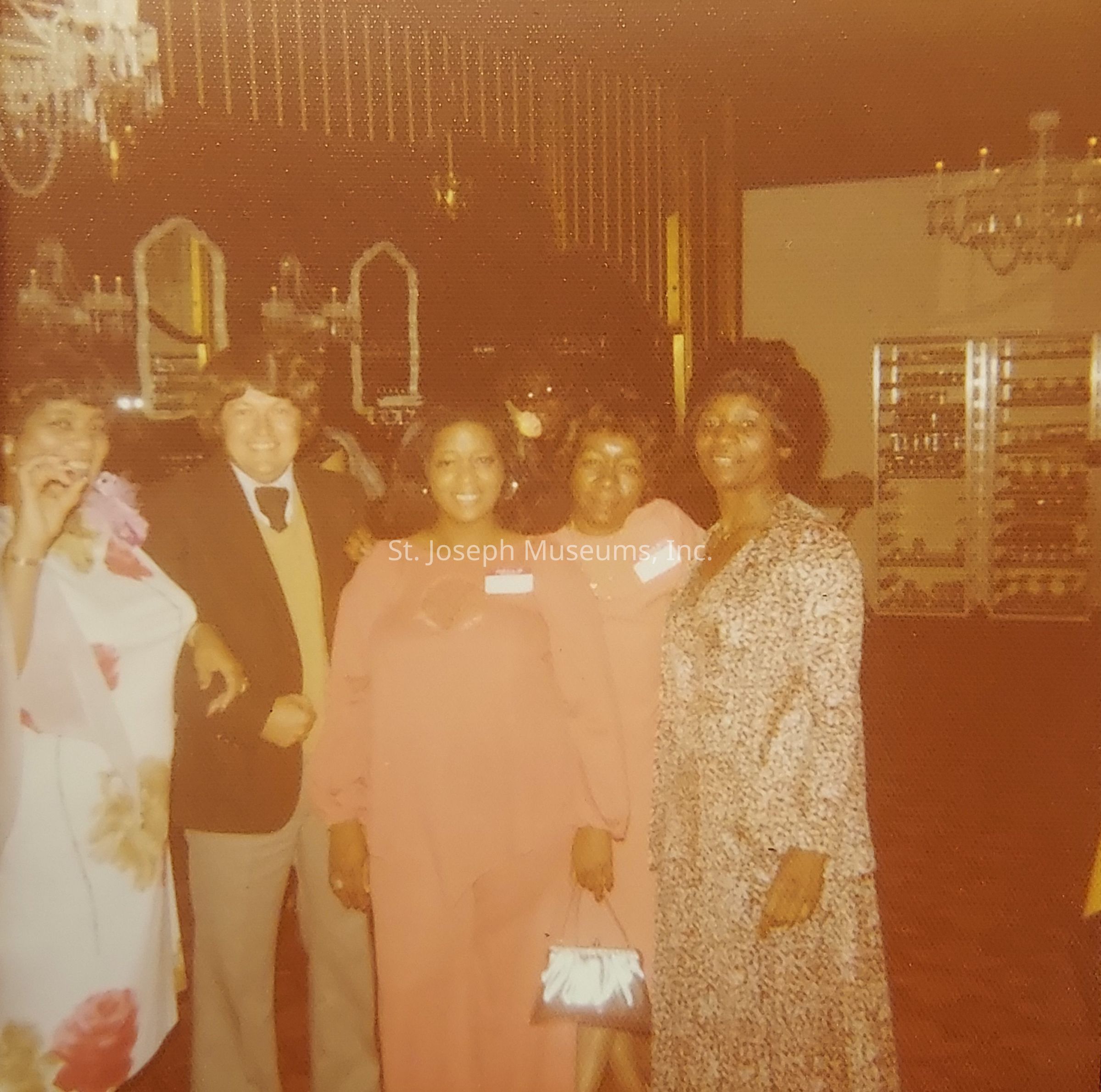 The image depicts a group of five people standing closely in what appears to be an elegant room, possibly a ballroom or banquet hall, adorned with large mirrors and chandeliers hanging from the ceiling, creating a sophisticated atmosphere. Three women in the center and one on the far right are dressed in elegant dresses, each wearing name tags, suggesting they might be attending a formal event or gathering. The man second from the left is wearing a suit with a tie, adding to the formal ambiance of the occasion. The person on the far left is holding a drink, indicating a festive or celebratory context. The room's decor, with its warm lighting and mirrors, contributes to the formal and exquisite setting, enhancing the overall sophisticated and celebratory atmosphere of the occasion captured in the photo.