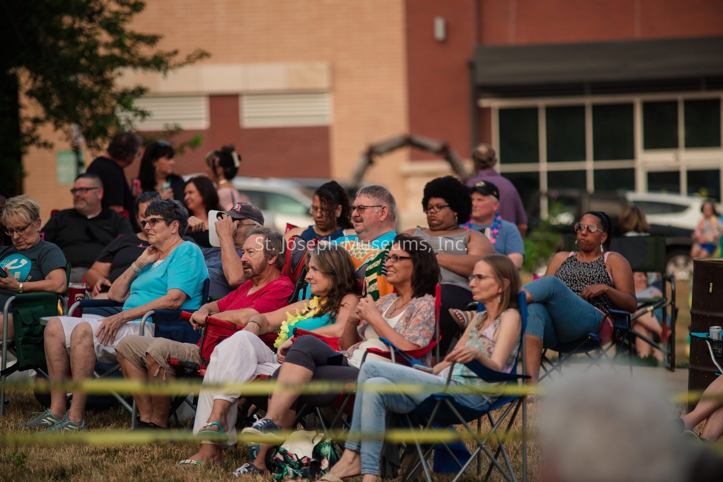 The image depicts a diverse group of people seated in folding chairs outdoors, seemingly attending an event or gathering on a grassy area. The scene is set against a backdrop of brick buildings, suggesting it's in an urban or community space. The attendees appear relaxed, engaging with the event in front of them, though none of the onlookers display overt excitement, maintaining a casual demeanor. Some are wearing sunglasses, indicating sunny weather, while others hold beverages, adding to the leisurely atmosphere. The foreground features yellow tape, possibly marking off the area. In the background, additional people are partially visible, indicating a larger crowd. The overall ambiance is calm and communal, with individuals enjoying a shared moment in a simple, open-air setting, perhaps taking in a performance or presentation. The variety in attire and expressions reflects a mix of personalities, highlighting the inclusive nature of the gathering.