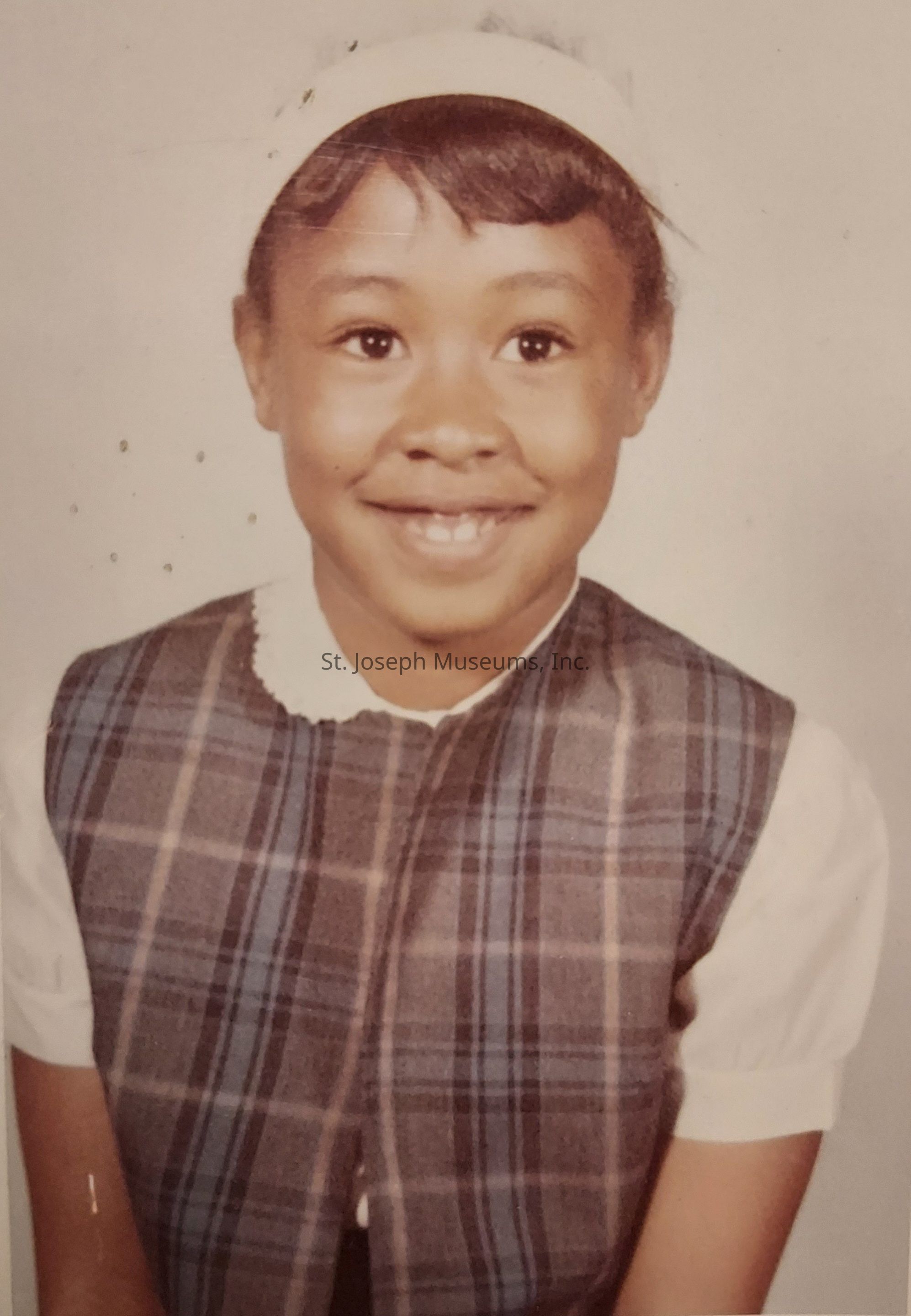 The image depicts a young girl smiling warmly at the camera. She is wearing a plaid dress with a white blouse underneath, which features a scalloped collar. Her hair is styled neatly, with bangs framing her forehead, complemented by a white headband. The background is plain and neutral, drawing focus to her cheerful expression. The photograph has a vintage feel, suggesting it might have been taken several decades ago, capturing the innocence and happiness of childhood.