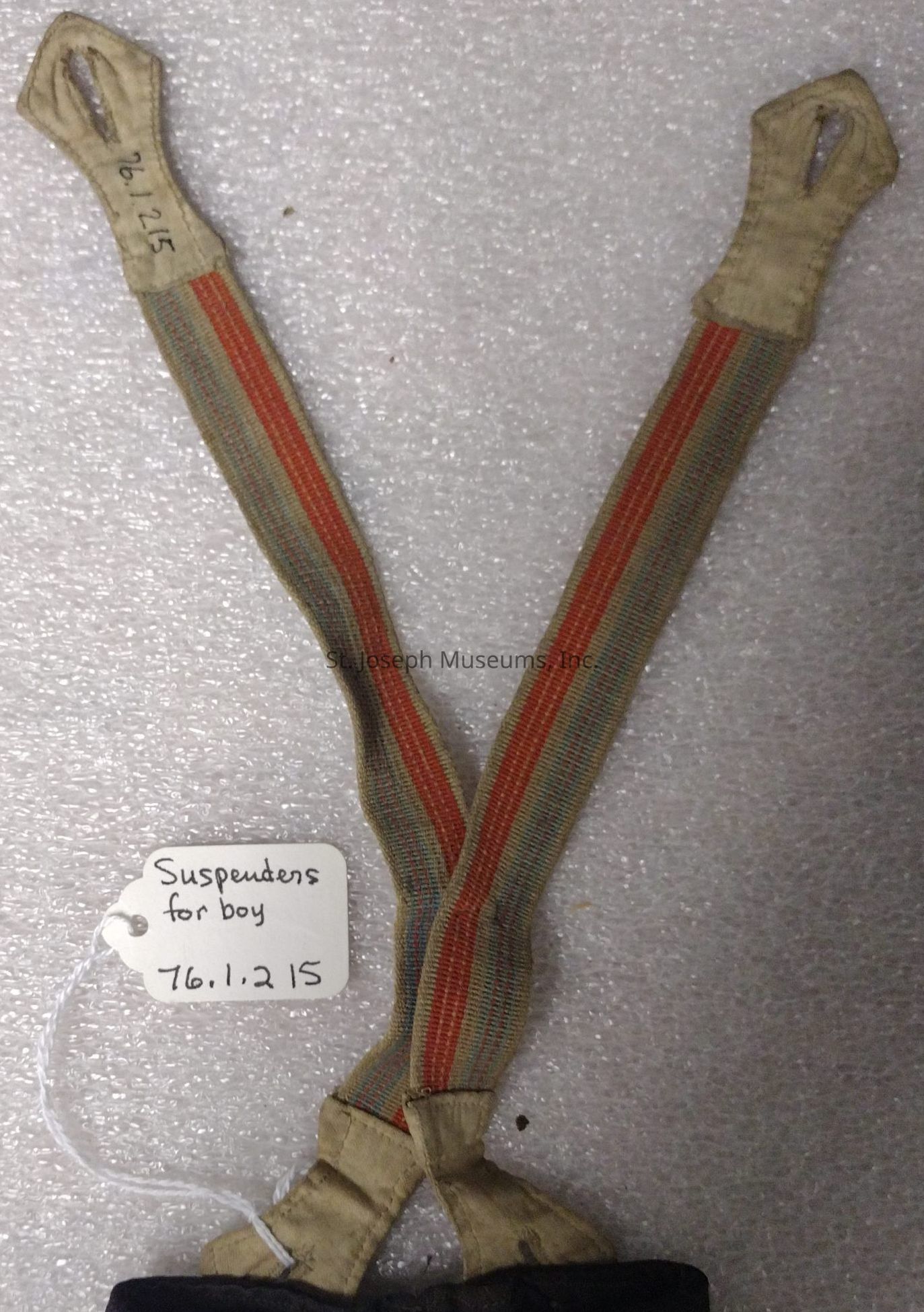 The image shows a pair of vintage suspenders designed for a boy, featuring an earthy tone with thin, colorful stripes running along the fabric, attached to two beige leather fasteners with buttonholes at the ends, and a handwritten label tied with string that reads "Suspenders for boy 76.1.2 15," against a textured, neutral background that provides a simple, yet detailed backdrop, highlighting the suspenders' craftsmanship and potentially historic value, while offering a glimpse into a bygone era of clothing accessories, possibly invoking a sense of nostalgia or appreciation for traditional apparel and accessory designs, and making it suitable for display in a museum or a collection specializing in historical garments for its visual appeal and apparent significance, thus allowing observers to reflect on changes in clothing fashion, utility, and the evolution of practical garment accessories over the years, complete with tags that might aid in cataloging and preserving historical artifacts or exhibits.