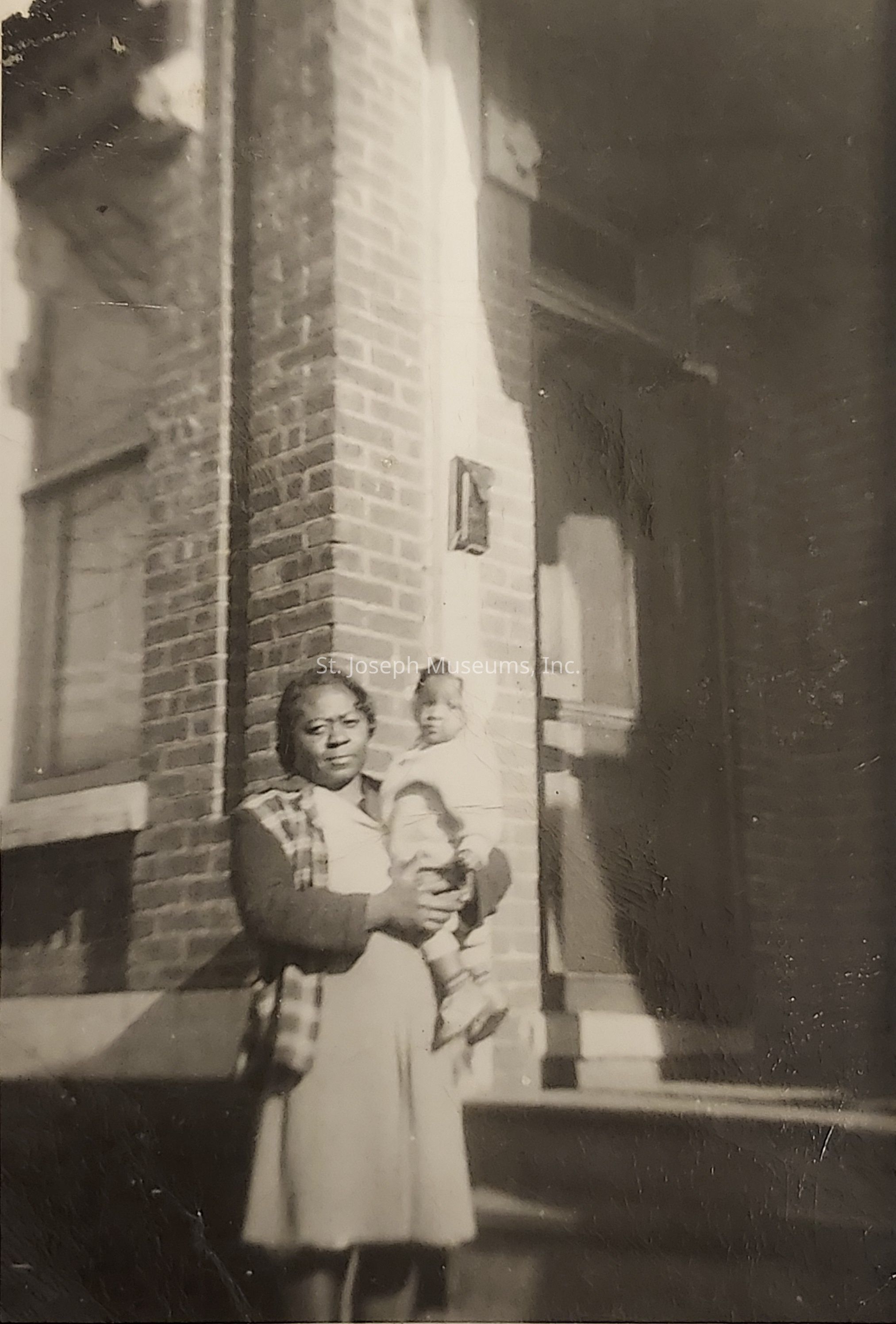 The vintage black and white photograph captures a woman standing in front of a brick building, holding a young child in her arms. She is wearing a dress and a cardigan, and the child is dressed warmly, wearing what appears to be a coat. The background features large windows and a doorway, indicating the scene is set in an urban environment, possibly from the mid-20th century. The expressions on their faces suggest a candid moment, and the overall tone reflects a sense of nostalgia and family connection. Shadows cast by the sunlight provide depth and highlight the texture of the building's brickwork.