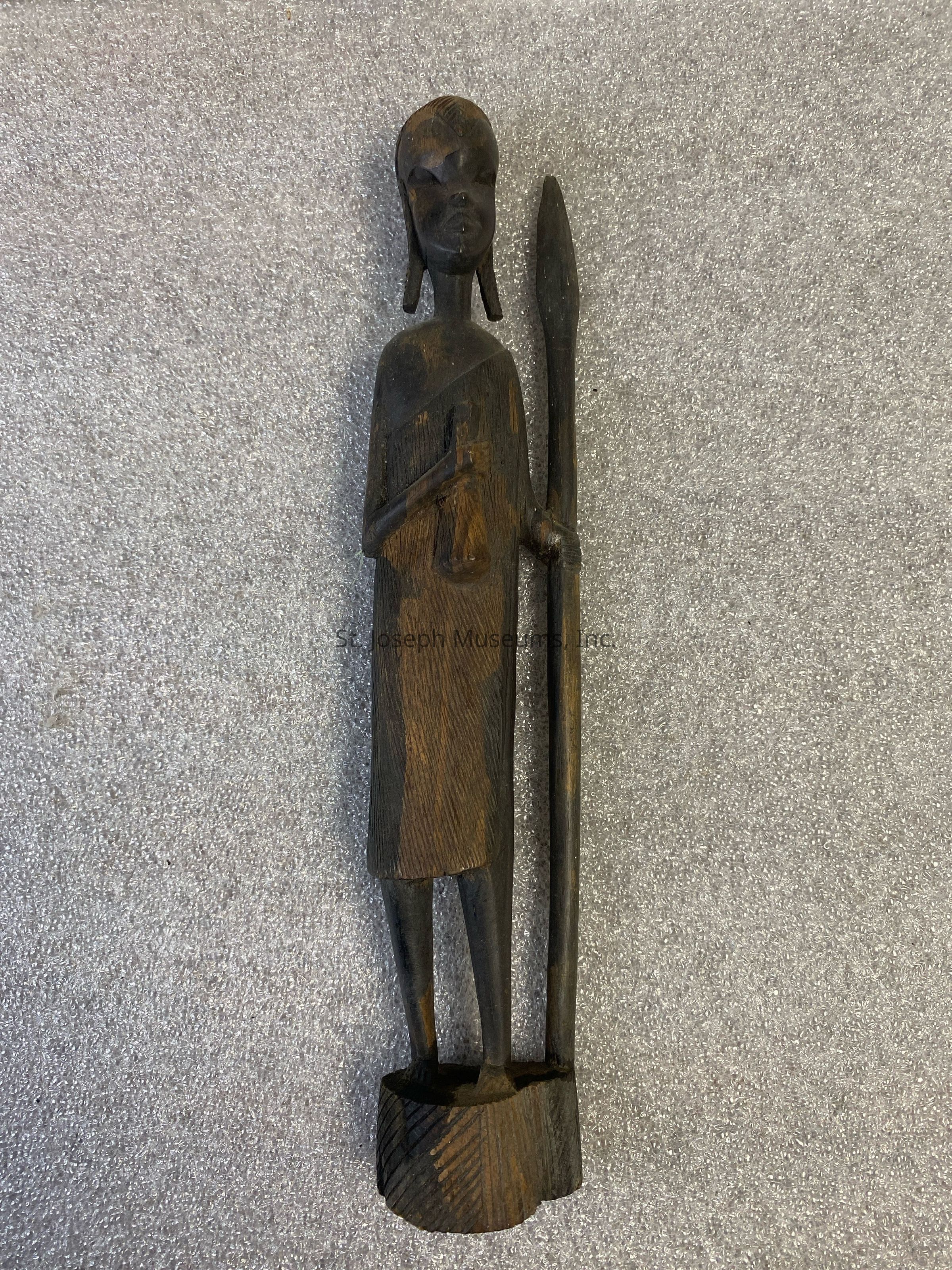 The image showcases a skillfully carved wooden sculpture depicting a standing figure with a serene expression, adorned in traditional attire, and holding a tall, slender spear-like object in one hand while the other hand rests calmly by their side. The intricate details on the clothing and headdress highlight the craftsmanship, and the dark wood gives the piece a rustic, timeless appearance, creating an elegant contrast against the textured gray background.
