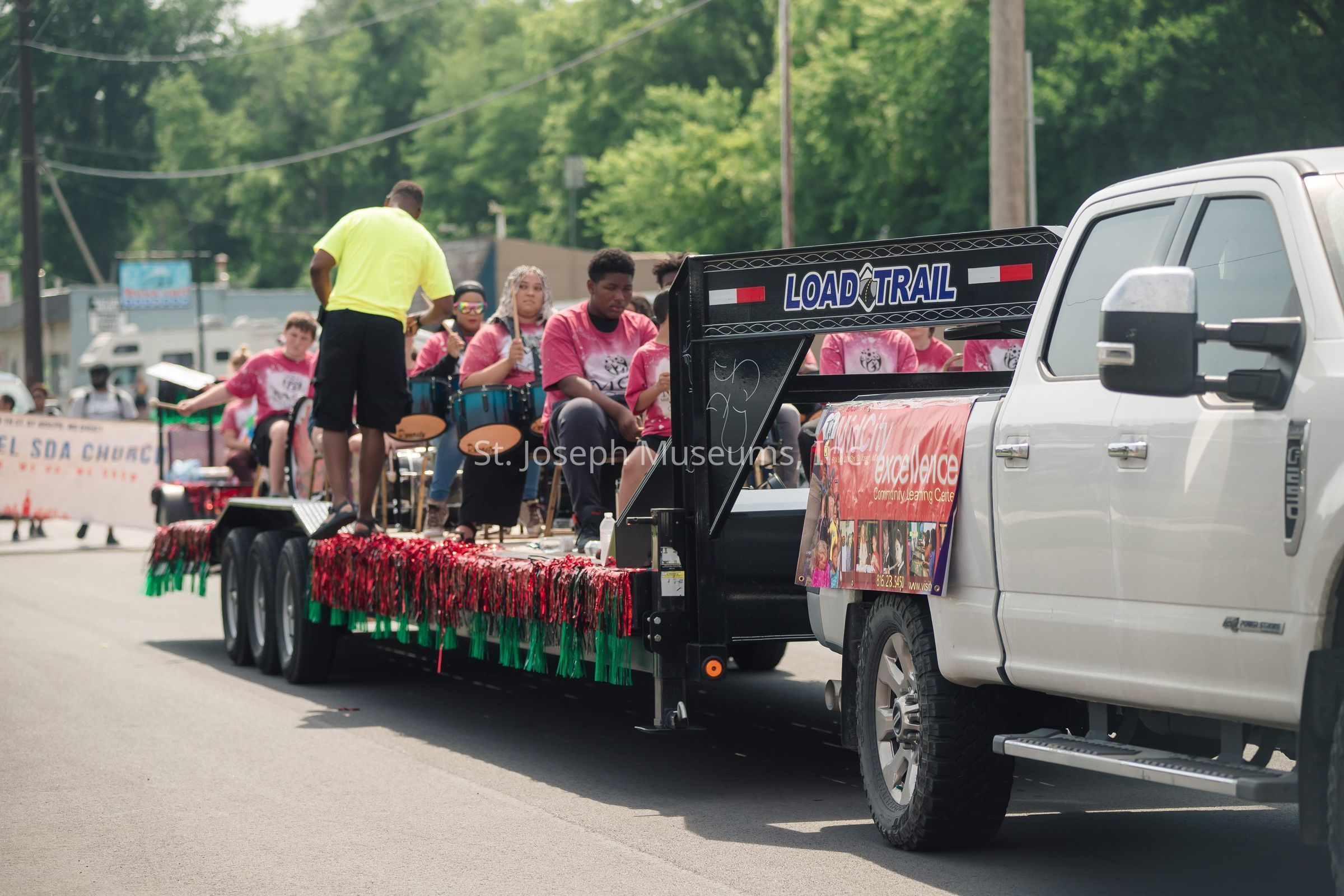 In the image, a large white pickup truck is towing a decorated trailer along a street, participating in what appears to be a parade. The trailer is adorned with festive red and green fringe, and several people, mostly dressed in matching pink T-shirts, are seated on it, some of whom are playing drums under a sunlit sky. A person in a bright yellow shirt is standing on the trailer, overseeing the group. A sign on the side of the truck reads "Twin City Excel-erate Community Learning Center," indicating the affiliations of the group. Behind them, a banner for a church is visible, further suggesting the community-focused nature of the event. The background is filled with lush green trees and various buildings, adding to the lively and cheerful atmosphere of the scene.