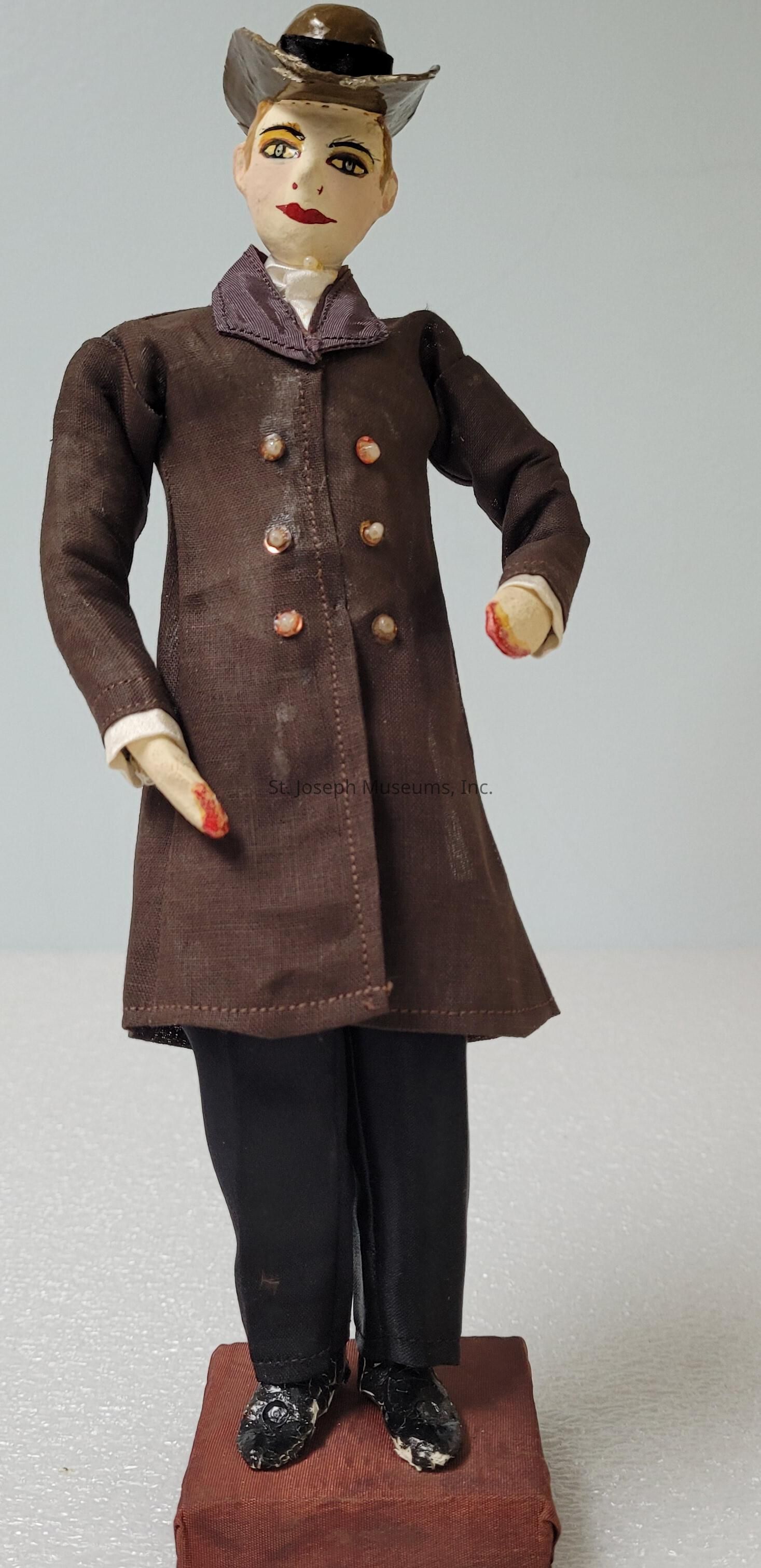 The image depicts a handcrafted doll dressed in vintage clothing, standing on a small platform. The doll is adorned in a brown coat with multiple buttons, paired with dark trousers. Its face is painted with expressive features, including arched eyebrows and red lips, and it wears a distinctive hat that complements its ensemble. The doll's hands are positioned gracefully, with slight wear evident on the fingers, indicating its aged and possibly cherished nature. Its shoes are black and appear textured, matching the overall antique style. The background is neutral, highlighting the doll's intricate details.