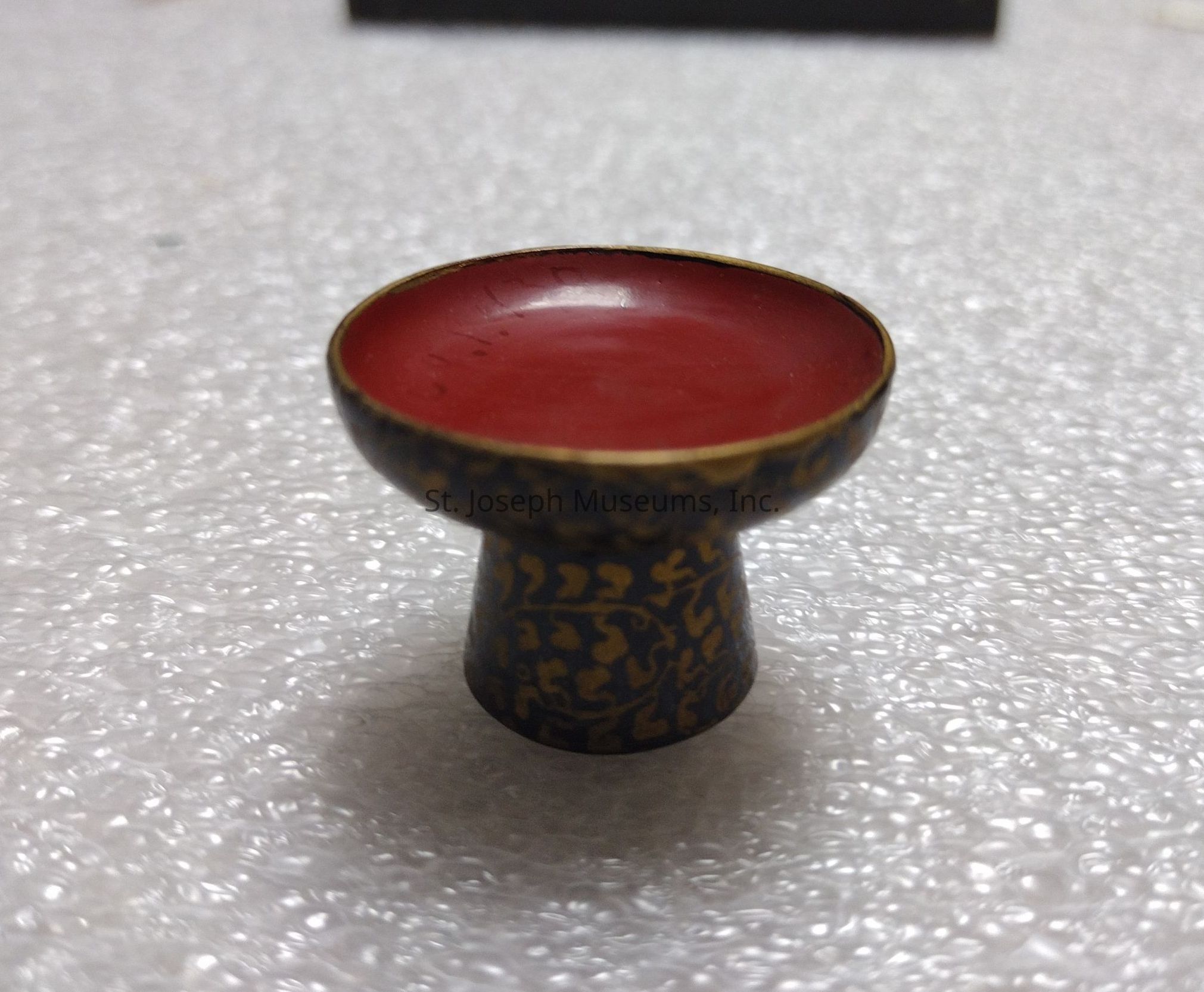 The image depicts a small, ornate cup or bowl with a wide, shallow, red interior and a short, sturdy base, possibly made of metal, with intricate gold patterns etched onto its dark exterior. The object is placed on a textured, bubble-like surface, which appears to be a protective wrapping material. The contrast between the cup's rich red interior and the gold-accented, dark exterior underlines its decorative and possibly cultural significance, suggesting it may be a ceremonial or artistic piece. The shallow design of the bowl, along with its decorative exterior patterns, highlights its aesthetic purpose. It's likely designed for display or specific ceremonial use rather than for practical utility.