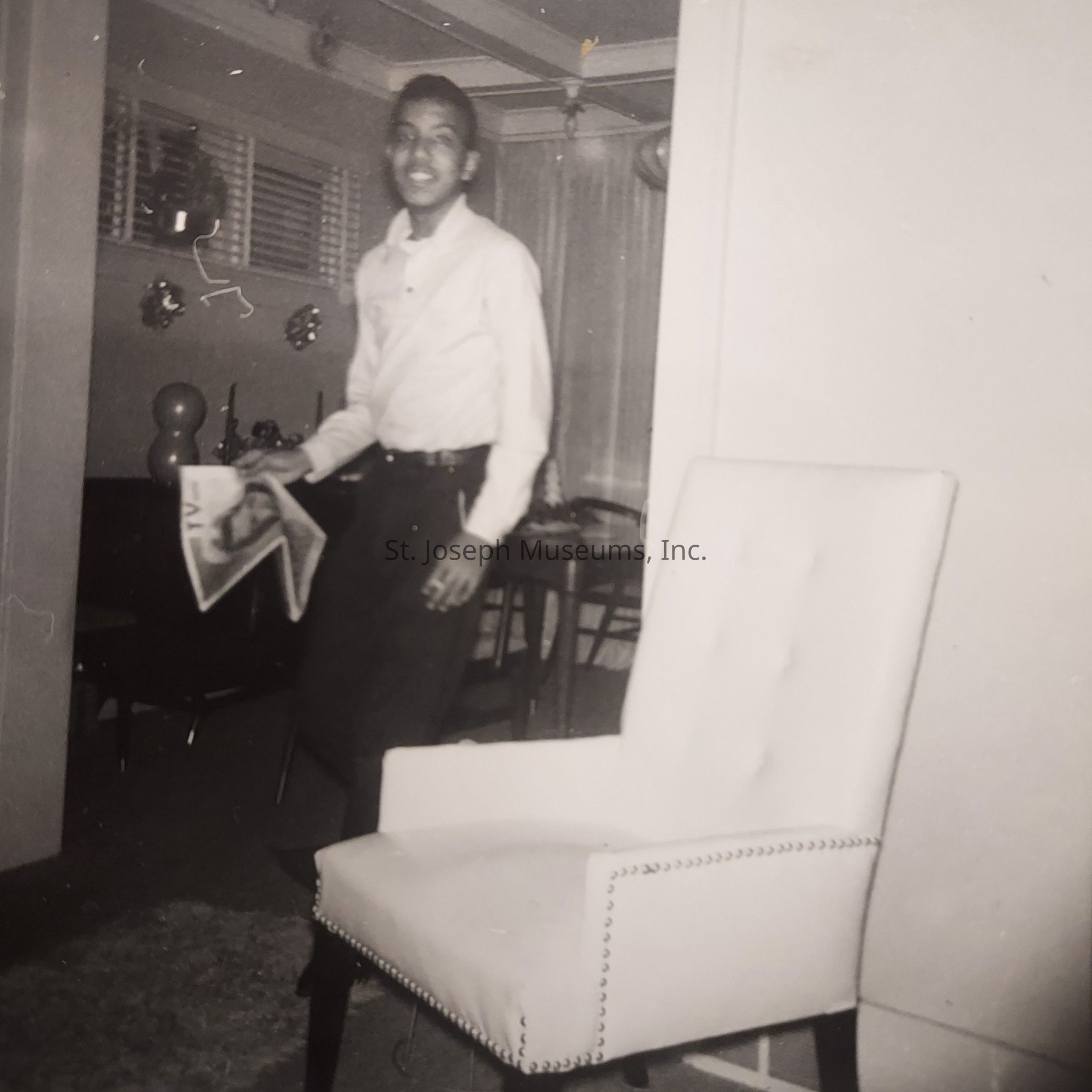 In this black-and-white photograph, a smiling man is captured mid-motion, holding a newspaper in his hand as he stands in a warmly furnished living room. The room features a comfortable white chair with studded detailing in the foreground, and in the background, there's a dining area with chairs and a table visible. The walls are adorned with small decorative items, possibly photographs or ornaments, enhancing the homely atmosphere. The man is dressed in a light-colored shirt tucked into dark trousers, embodying a casual yet neat style. Soft light filters into the room through horizontal blinds, creating a cozy ambiance that suggests a moment of leisure and everyday life captured in time. The overall composition exudes a sense of contentment and nostalgia, inviting the viewer to imagine the story behind this candid moment.