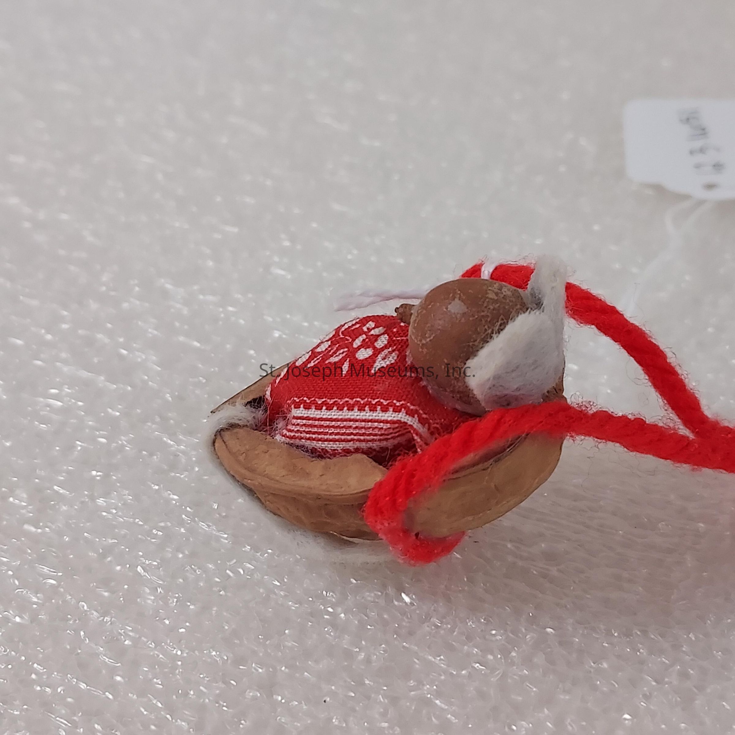 The image shows a small, creatively crafted figurine of a baby wrapped snugly in a red cloth with white patterns, nestled inside a half walnut shell. The walnut shell cradles the baby, giving it a cozy appearance. A tiny, round, brown bead forms the baby's head, with a white felt piece as the hat or hair. A red string is attached, possibly for hanging as an ornament. This miniature scene is placed against a textured, cushioned white background, creating a delightful contrast and emphasizing the intricate details and the careful craftsmanship of the tiny creation, making it a charming and imaginative representation.