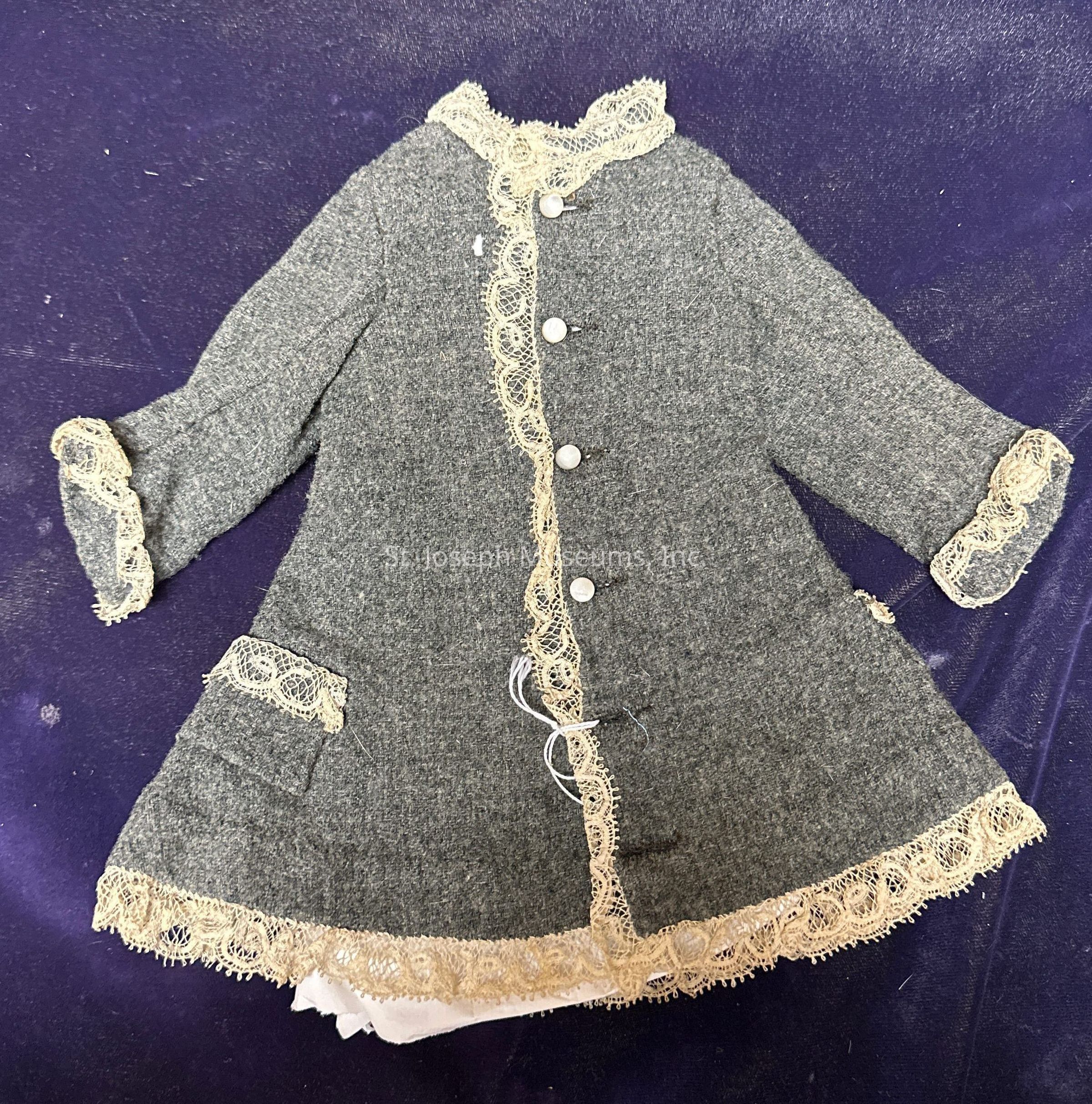 The image shows a vintage-style coat laid out on a deep purple background. This coat is made of a textured gray fabric and features decorative details that bring a touch of elegance to its design. Along its front, a row of four silver or metallic buttons fasten the coat securely. The edges, including the cuffs, collar, and bottom hem, are adorned with intricate cream-colored lace trim that adds a delicate, antique aesthetic. The coat also includes two small pockets on its front, bordered with the same elegant lace, enhancing its classic appeal. The long sleeves suggest that it is designed for cooler weather, while the overall design indicates it may be a piece intended for formal or classic attire. The positioning on the purple background highlights the coat’s subdued yet refined style, allowing its details to stand out clearly. The lining visible at the hem suggests added comfort and warmth. Overall, the coat embodies a blend of classic charm and vintage sophistication through its careful detailing and thoughtful design elements.