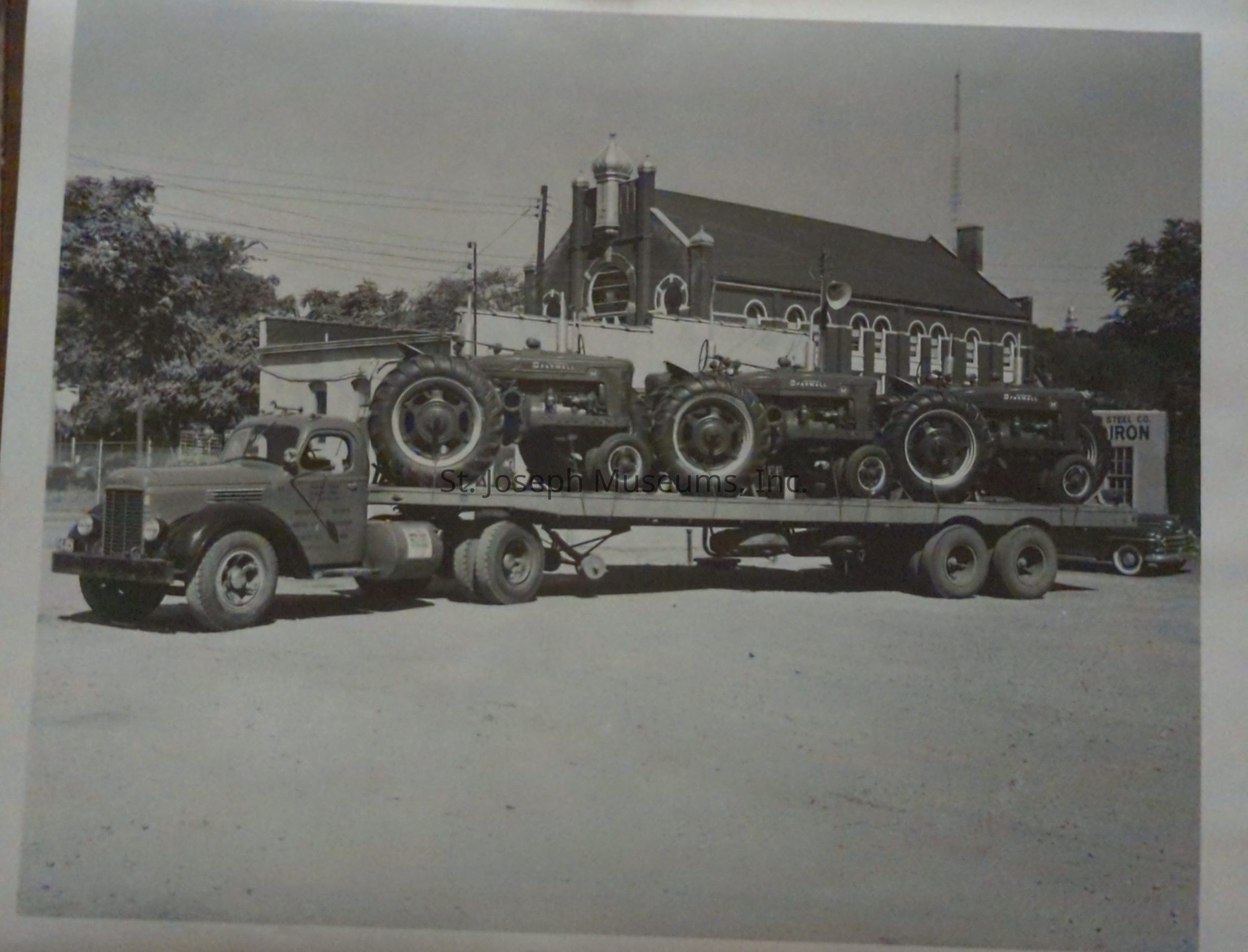 Black-and-white photograph of a vintage Crouch Bros. Co. truck is seen parked on South 7th Street, carrying a flatbed trailer loaded with three International Harvester Farmall tractors. A large building with distinctive architecture is visible in the background, along with a sign that reads "CAST IRON."