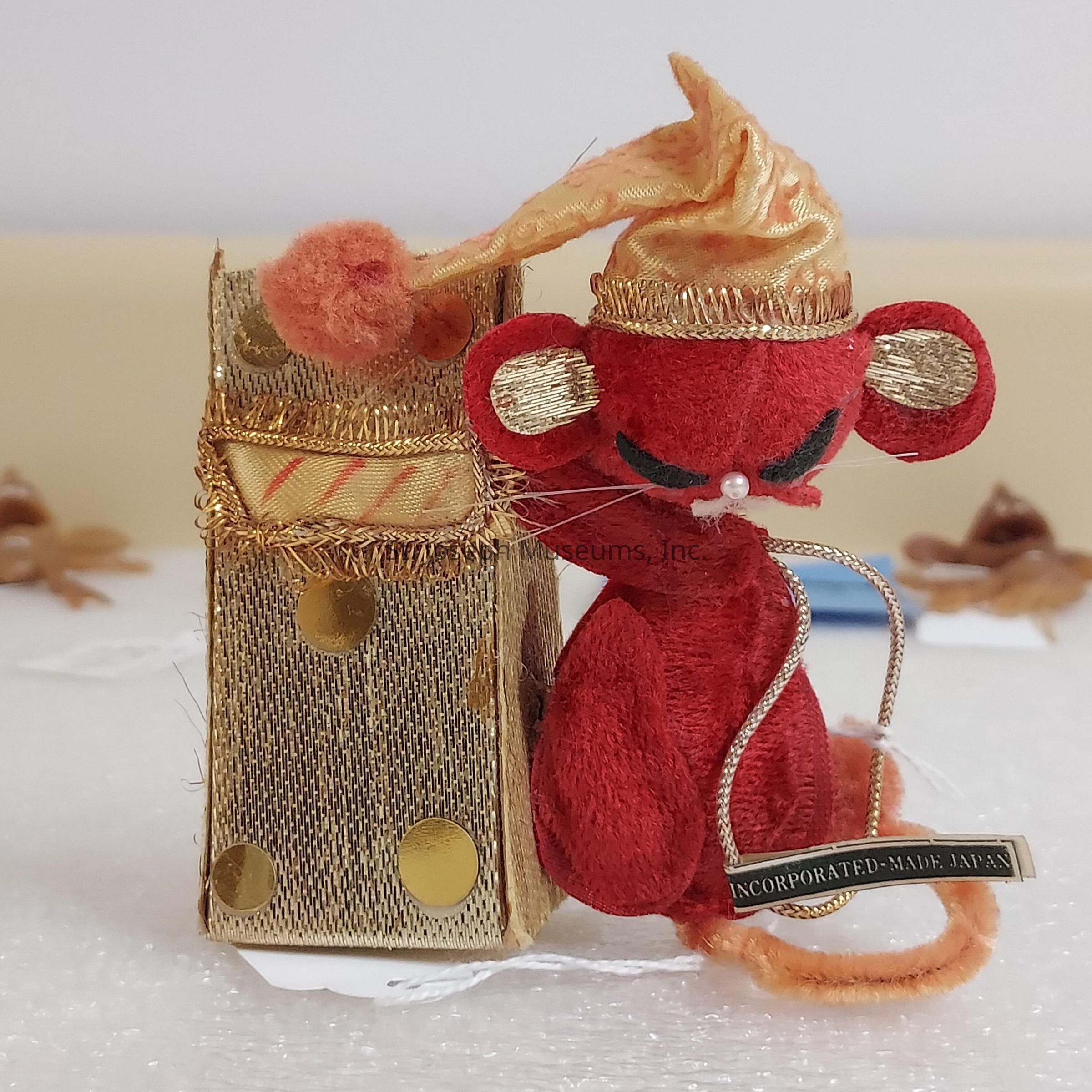 The image depicts a plush red mouse adorned in a festive hat, exuding a whimsical and charming appearance. The mouse, featuring closed eyes and a white whisker detail, stands beside a gold-toned, textured bag embellished with shiny polka dots. Both the mouse and the bag exhibit intricate details and craftsmanship, including gold stitching and design elements that enhance the overall visual appeal. At the mouse’s breast, a small label reads "Incorporated - Made in Japan," highlighting its manufactured origin. The background is softly blurred, with autumnal elements such as brown leaves, adding seasonal context and depth to the scene.