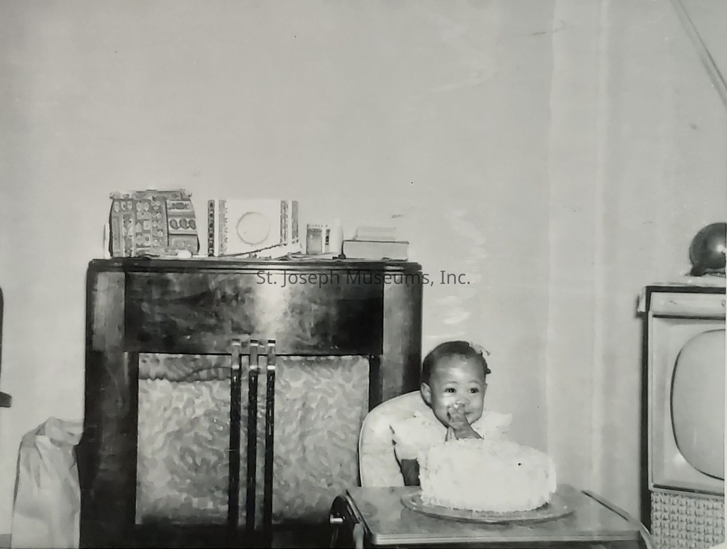 In a charming vintage setting, a young child is centered and caught joyfully engaged with a large, fluffy cake placed in front of them. The child, seated in a high chair, has a playful expression with a finger brought to the mouth, as if tasting the cake. The room, exuding a nostalgic ambiance, is adorned with furnishings typical of a mid-20th century home. On the left, a sturdy wooden cabinet sits with items arranged on top, including decorative boxes and possibly a radio. On the right is an old-style television, its rounded screen encased in a frame, adding to the retro feel. Despite the monochrome nature of the photo, the warmth and happiness of the moment are palpable. The child’s innocent curiosity and delight in the experience capture the essence of youthful joy and celebration, likely marking a significant occasion such as a birthday. This timeless image encapsulates the simplicity and charm of childhood in an era gone by, filled with love and familial affection.