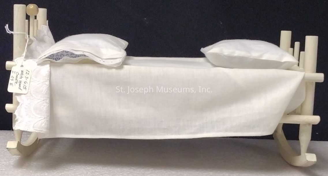 The image depicts a small wooden doll bed, featuring a cradle-like structure with a simple design and painted in a light color. The bed is adorned with a white sheet that drapes over its sides, and each of the two pillows is covered in matching white fabric. At one end, there is a lace-trimmed piece of cloth, adding a touch of elegance to the otherwise minimalist aesthetic. A handwritten tag is attached to the side, likely providing information about the object, but its details are not fully legible in the image. The dark background contrasts with the light-colored bed, highlighting its features.