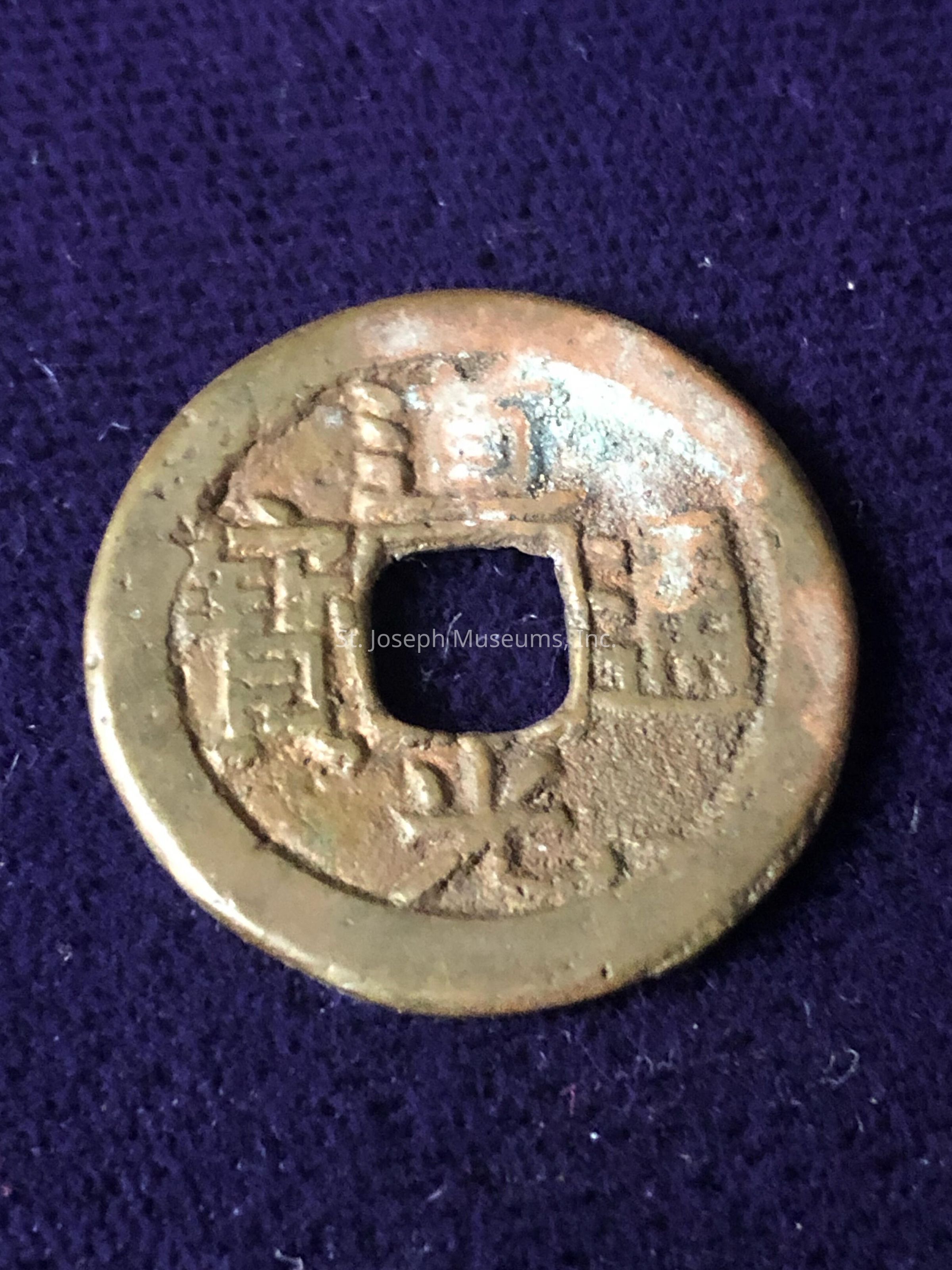 Coin A: The image features an ancient-looking, round coin made of metal, possibly bronze, with a square hole in the center, lying on a textured, deep purple surface. The coin showcases a patina indicative of age and wear, with inscriptions or characters etched around the square hole. The design is reminiscent of old Chinese coins, often used in ancient times for trade and currency. The surface of the coin appears slightly oxidized, highlighting a mix of green and brown hues. The intricate details of the markings suggest historical significance and possible cultural value, capturing a glimpse of past monetary systems.