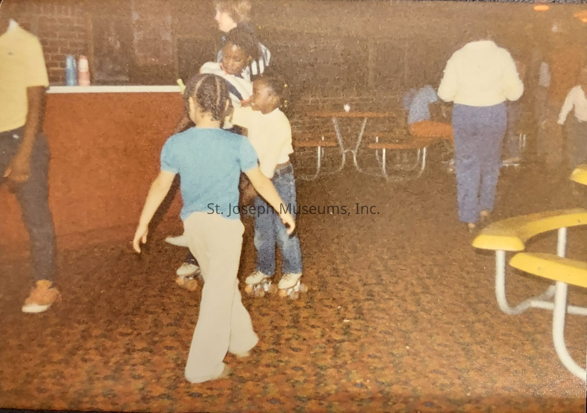 In the image, a group of children is roller skating inside an indoor rink with a vividly patterned carpet. The scene appears to be from the past, characterized by the style of their clothing and roller skates. One child in a blue top and light-colored pants is skating ambitiously, with another pair of children nearby, one wearing a yellow shirt and jeans. In the background, there are red walls, a counter with cups, and picnic-style seating with yellow benches, suggesting a location meant for gathering and fun. The environment exudes a nostalgic vibe, likely capturing a moment of joy and playfulness from a past era. The expressions and positions of the children indicate movement and interaction, contributing to the lively atmosphere of the roller rink.